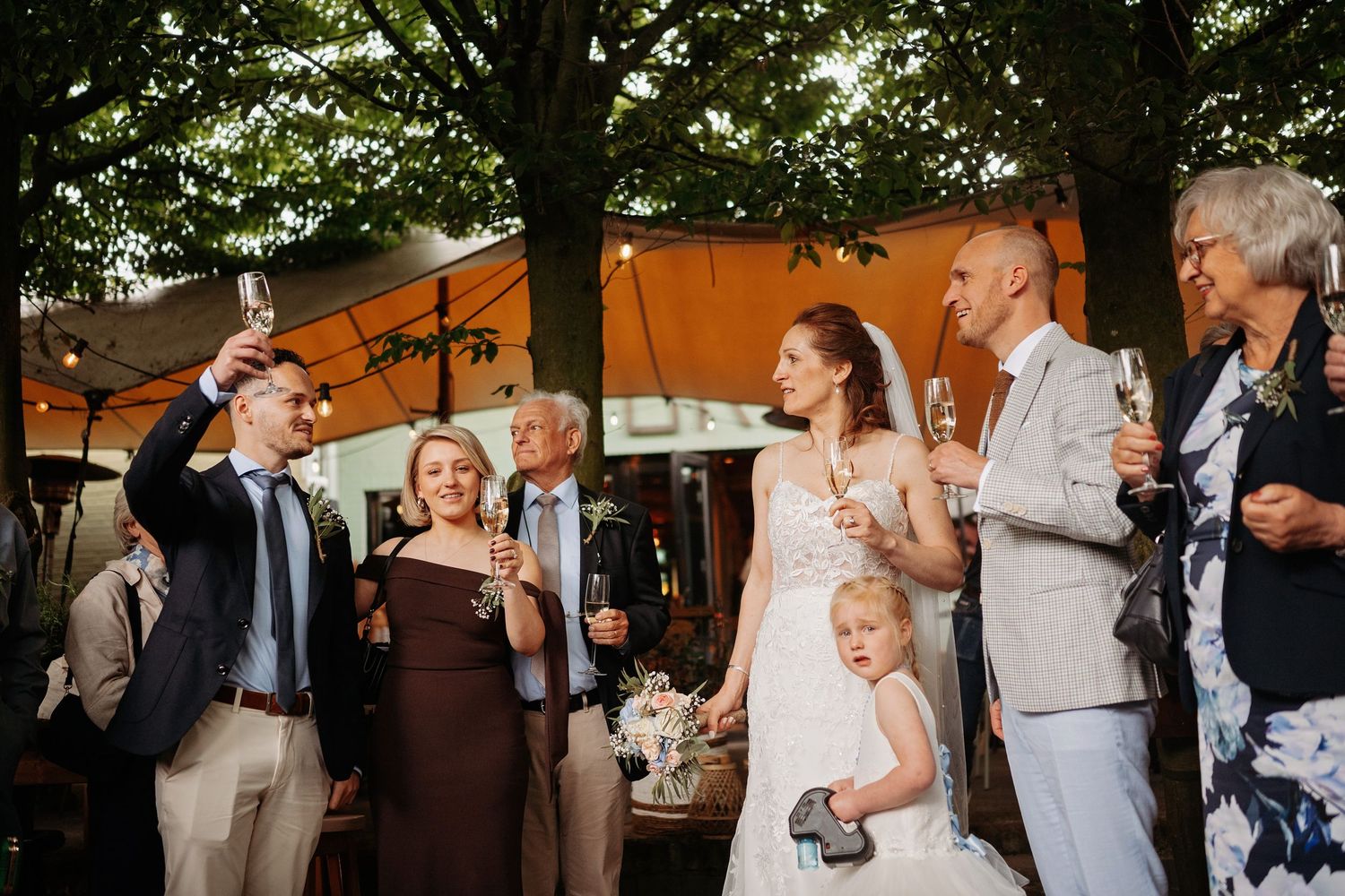 Wedding guests toasting with champagne outdoors under trees, bride and groom celebrating with family.
