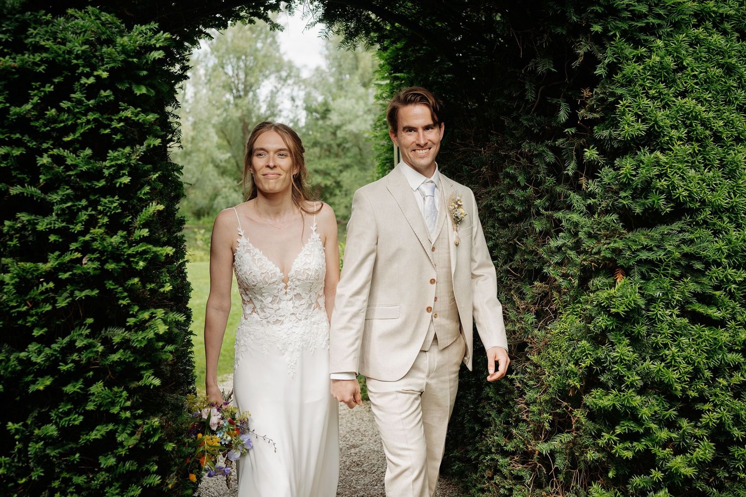 Bride and groom walk hand in hand through lush green hedgerow archway, bride in white gown, groom in beige suit.