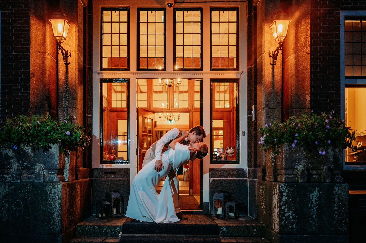 Groom dips bride in white gown for a romantic kiss outside an illuminated brick wedding venue at dusk.