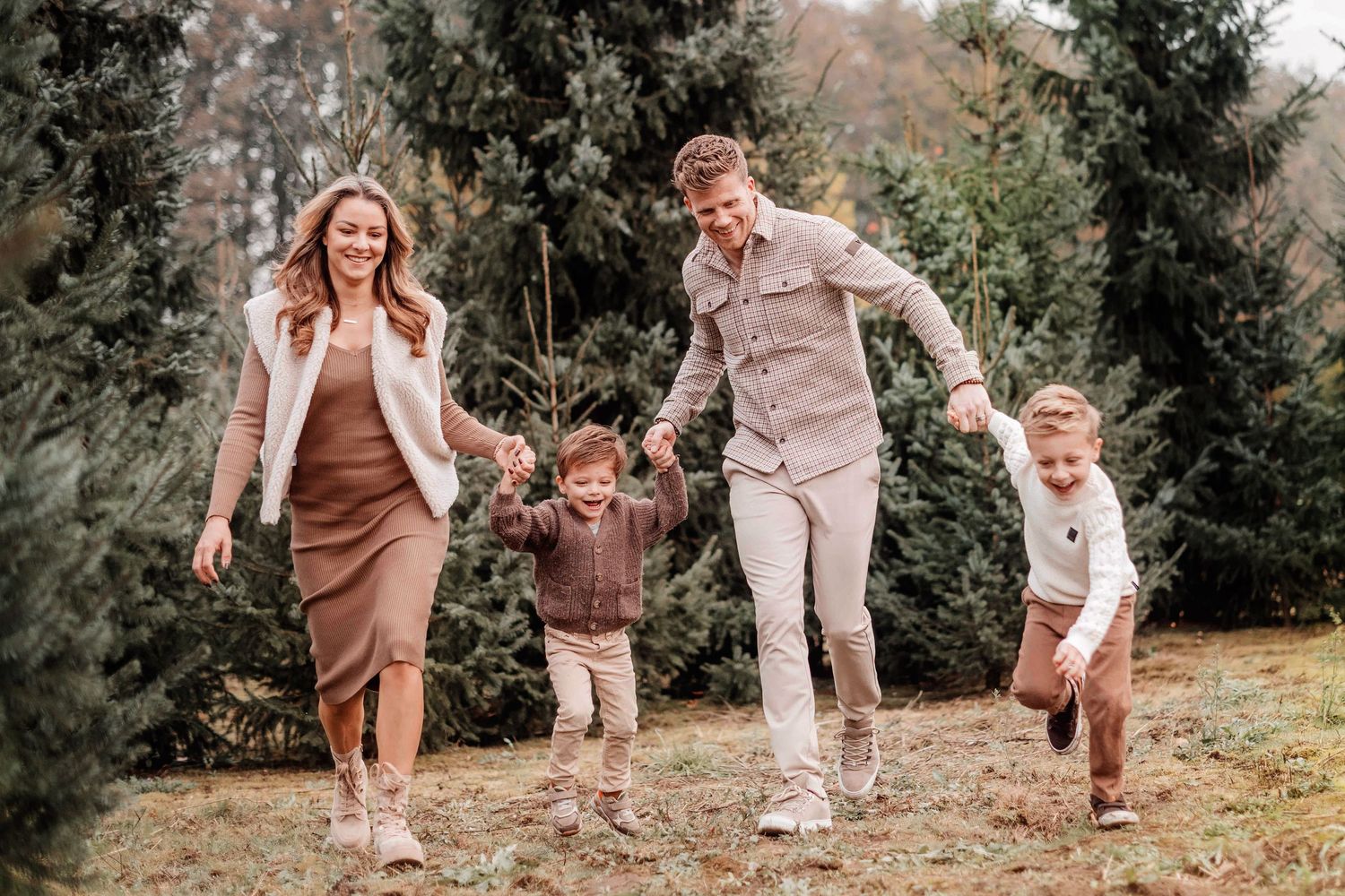 Happy family of four holding hands and running outdoors near evergreen trees in coordinated neutral fall outfits.