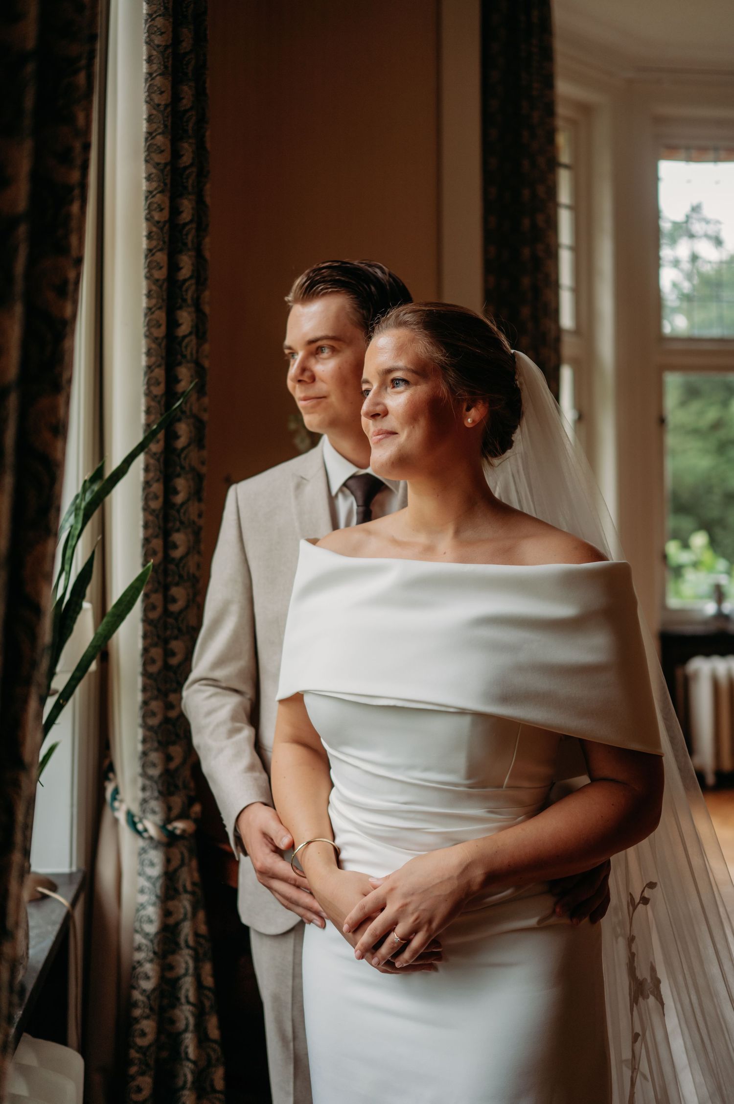 Bride in off-shoulder white gown and groom in gray suit share a tender moment by a sunlit window.