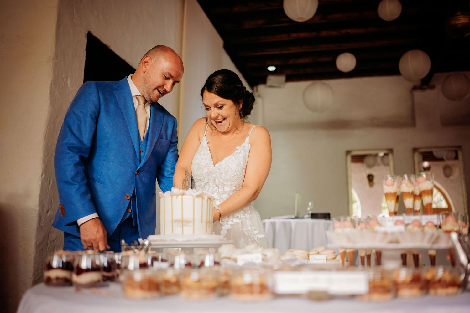 A bride and groom in blue suit cut their wedding cake at a dessert table display.