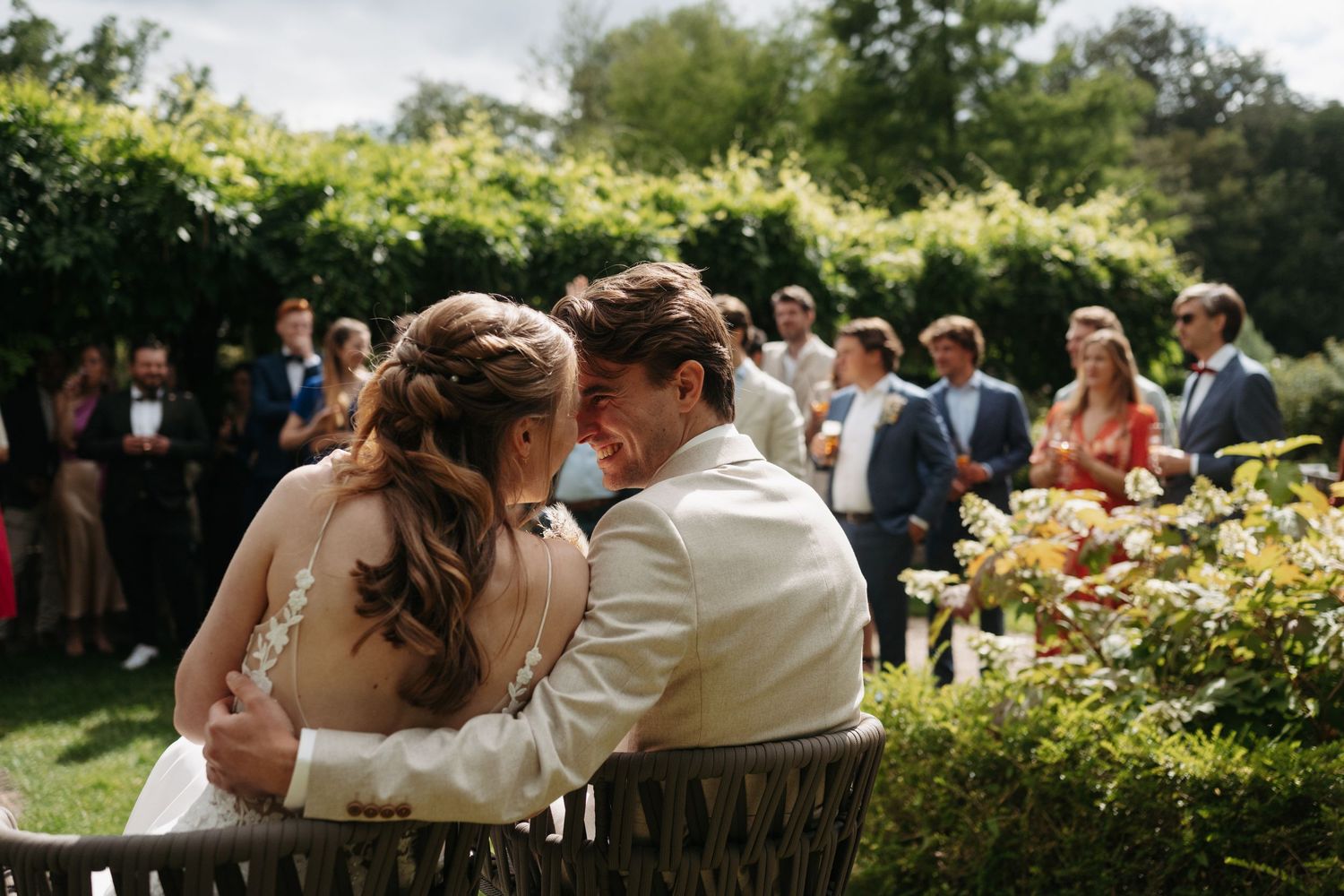 Bride and groom share an intimate moment seated outdoors at a garden wedding ceremony with guests in the background.
