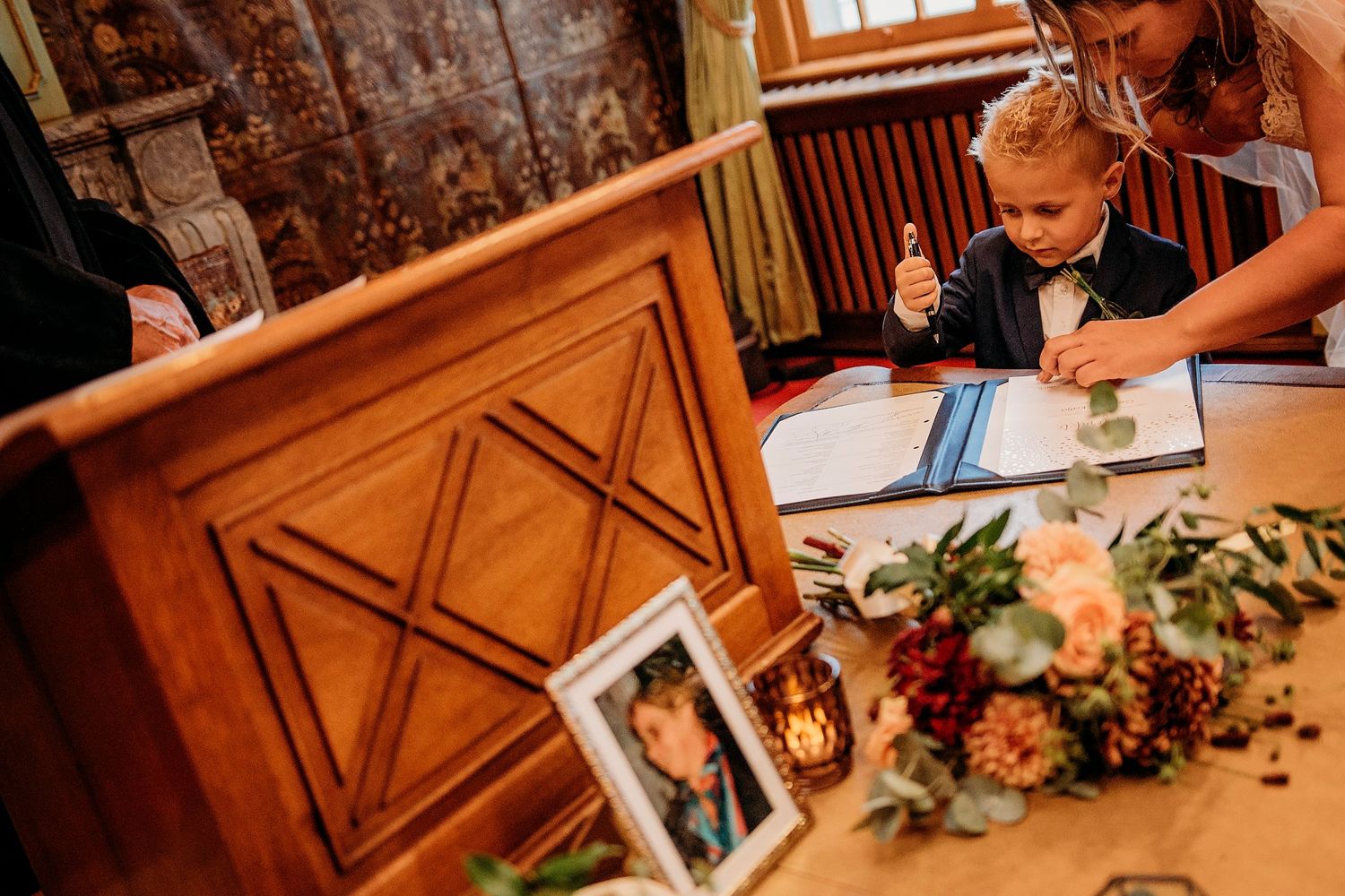 A young child signs a wedding registry book at a wooden table decorated with flowers and a framed photo during a ceremony.