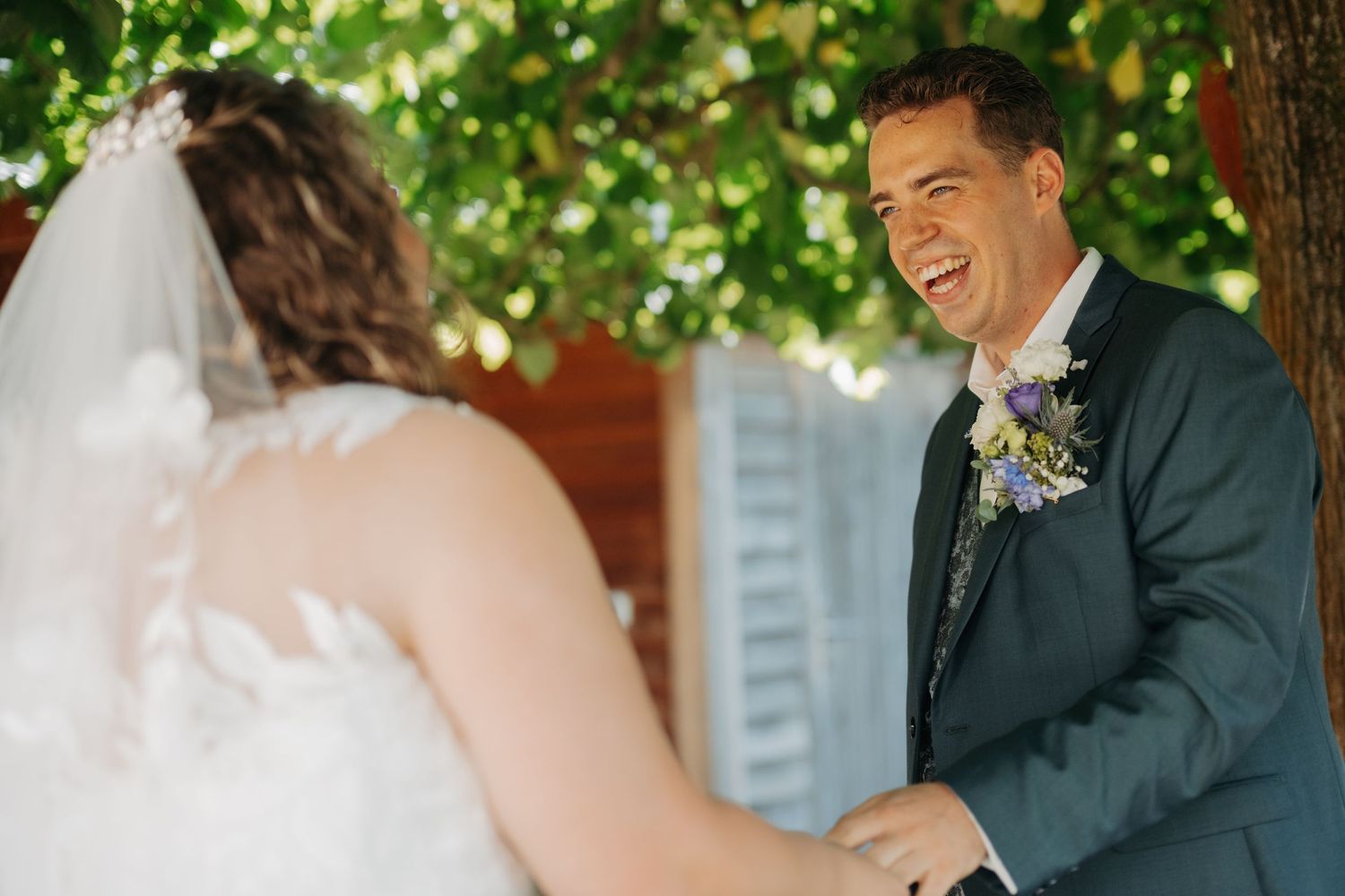 Groom in blue suit laughing joyfully during first look with bride in lace wedding dress outdoors.