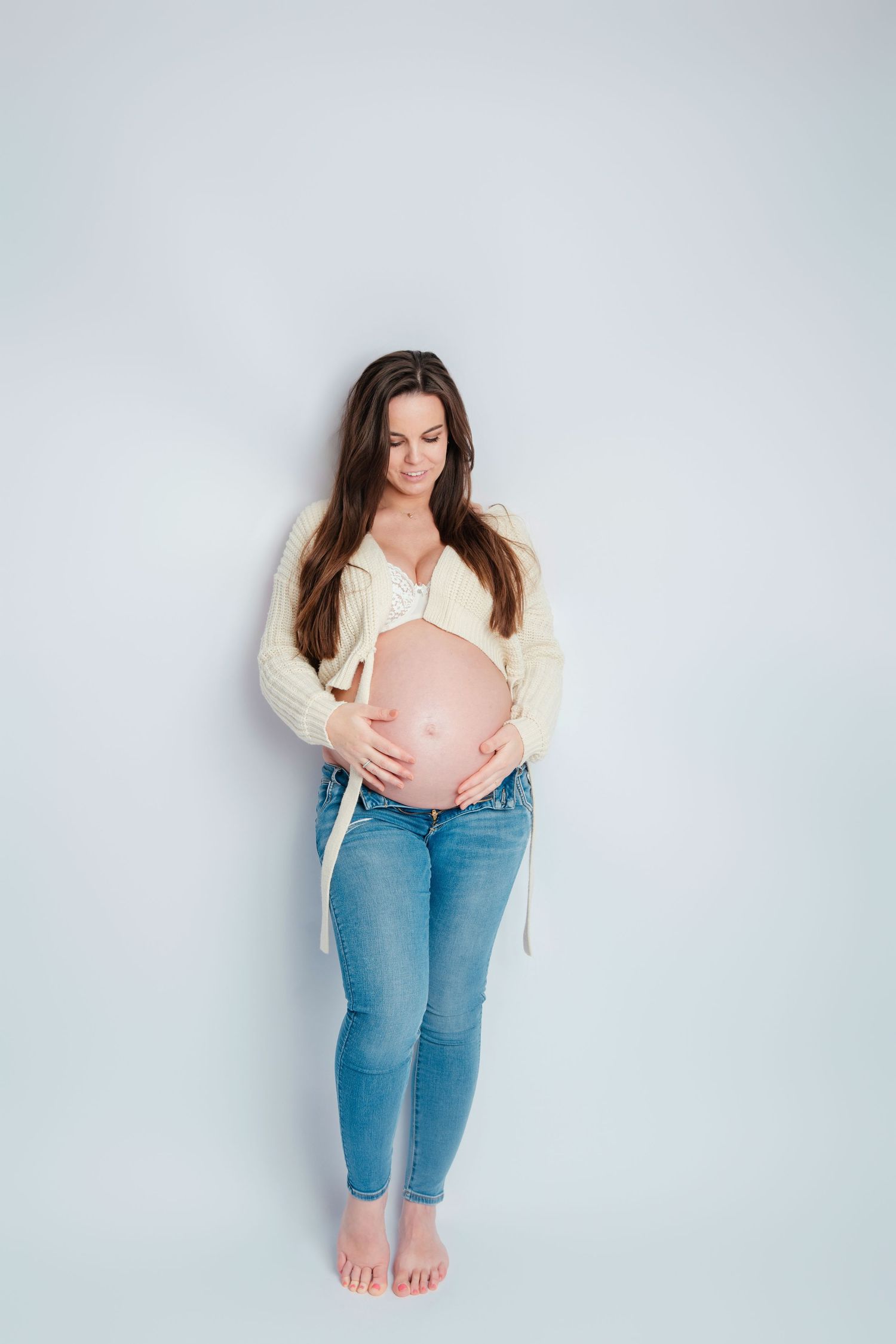 Toekomstige moeder poseert in crèmekleurige cardigan en jeans tegen witte muur, zwangerschapsfotografie Vught, Brabant.