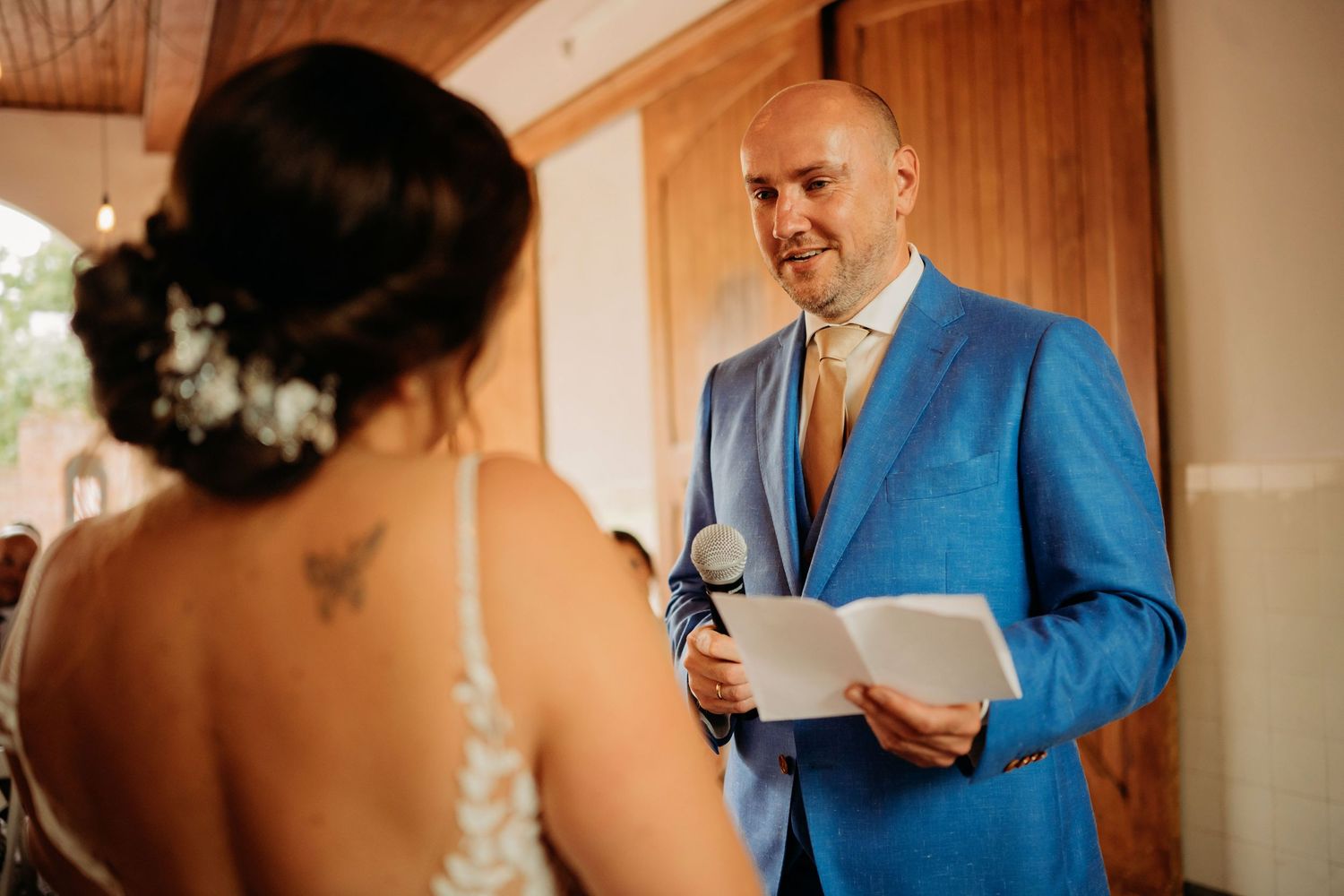 Groom in blue suit reads heartfelt vows from paper to bride during intimate indoor wedding ceremony.