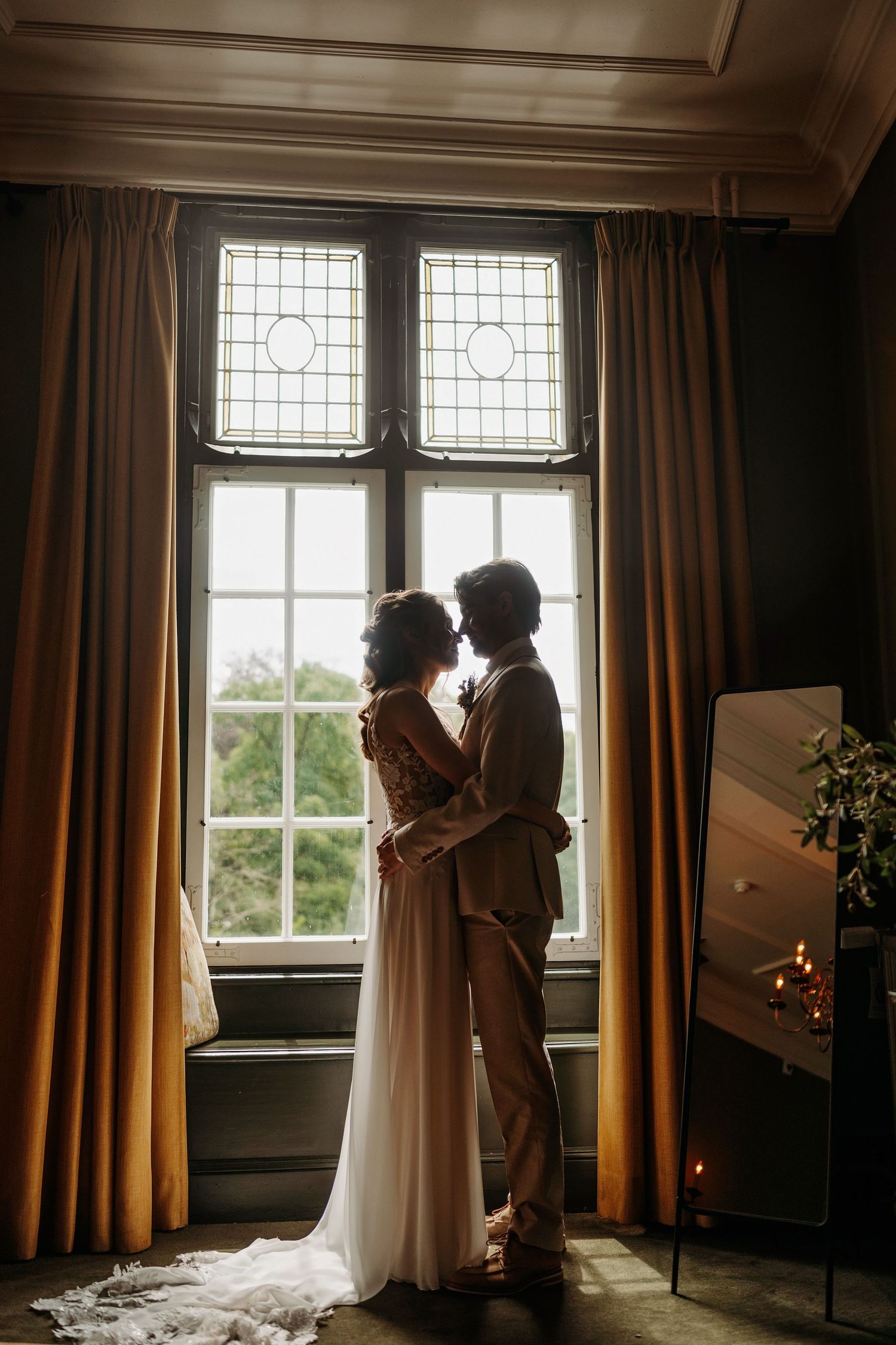 Couple silhouetted against a large leaded window in a dark elegant room, bride in flowing white gown embracing partner.