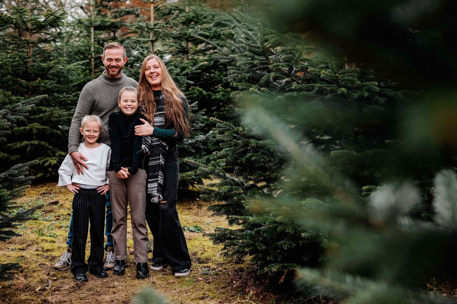 Family of four smiling together at a Christmas tree farm surrounded by lush green pine trees.