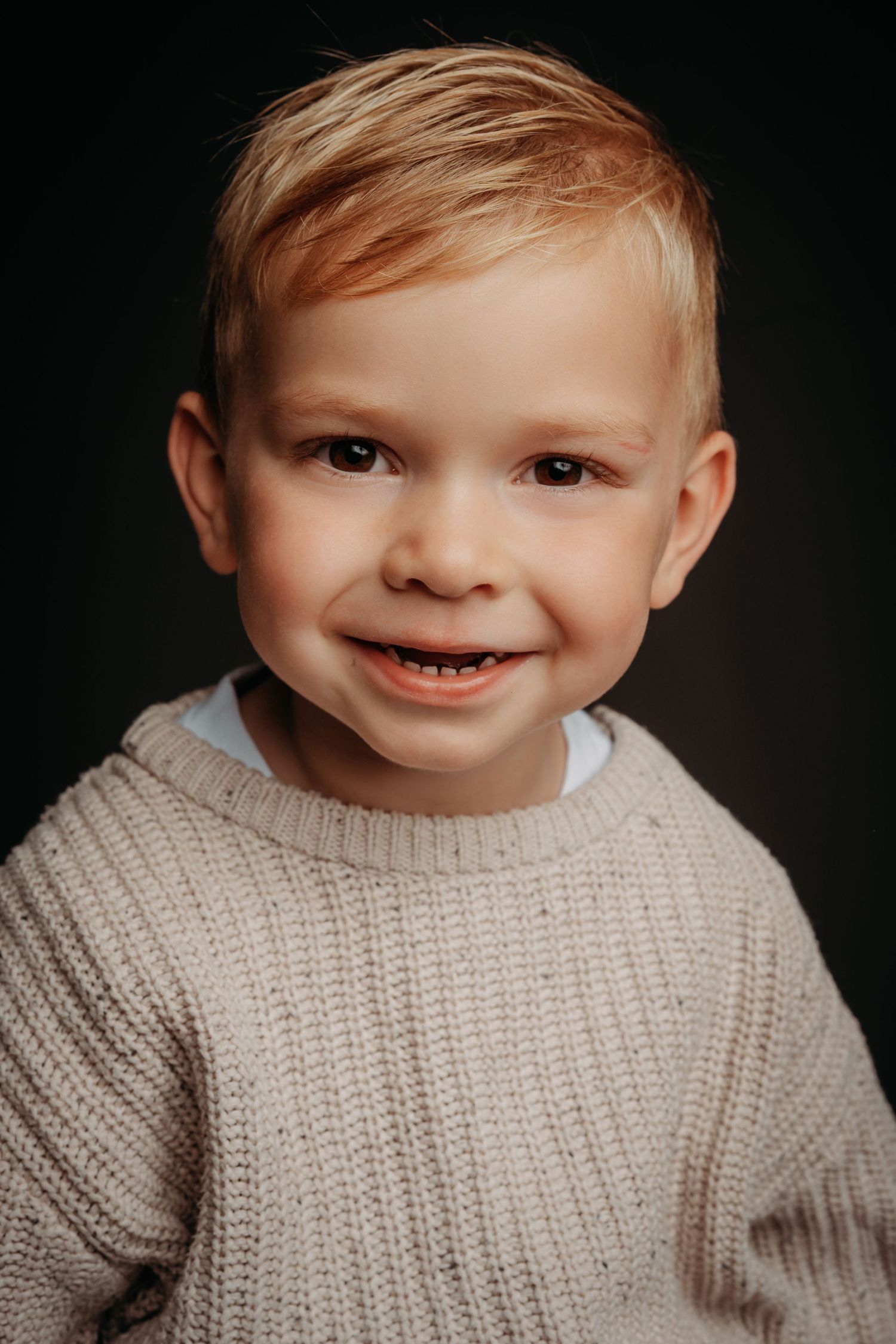 Smiling toddler boy with blonde hair wearing a beige knit sweater, portrait photo on dark background.