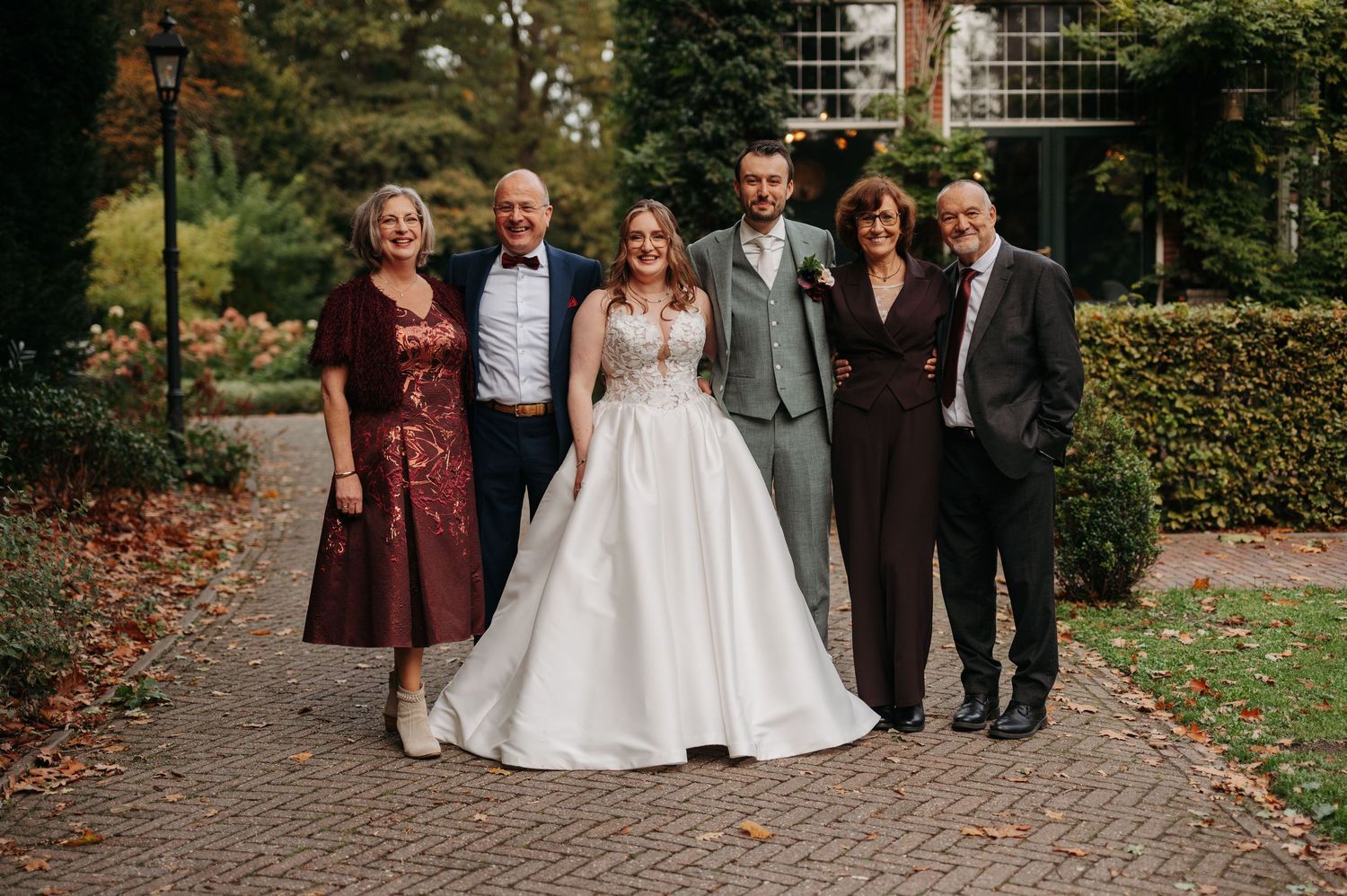 Bride in a white gown poses with six family members in formal wedding attire on a brick path beside a garden.