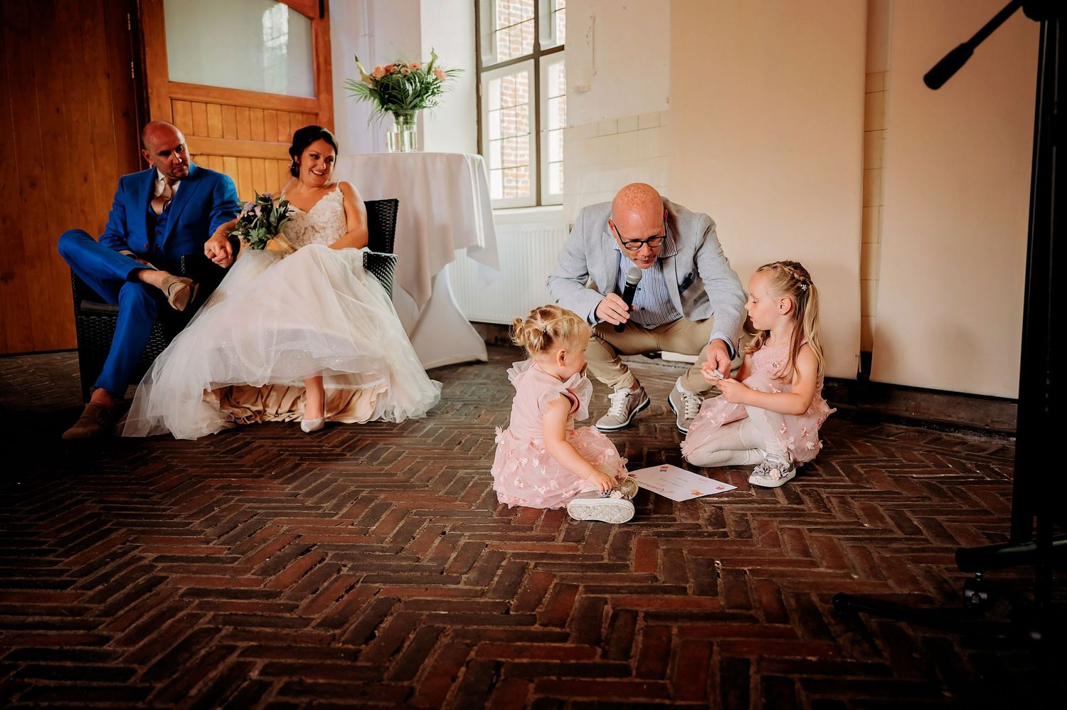 Bride and groom relax while a man entertains two young flower girls on a brick floor at a wedding venue.