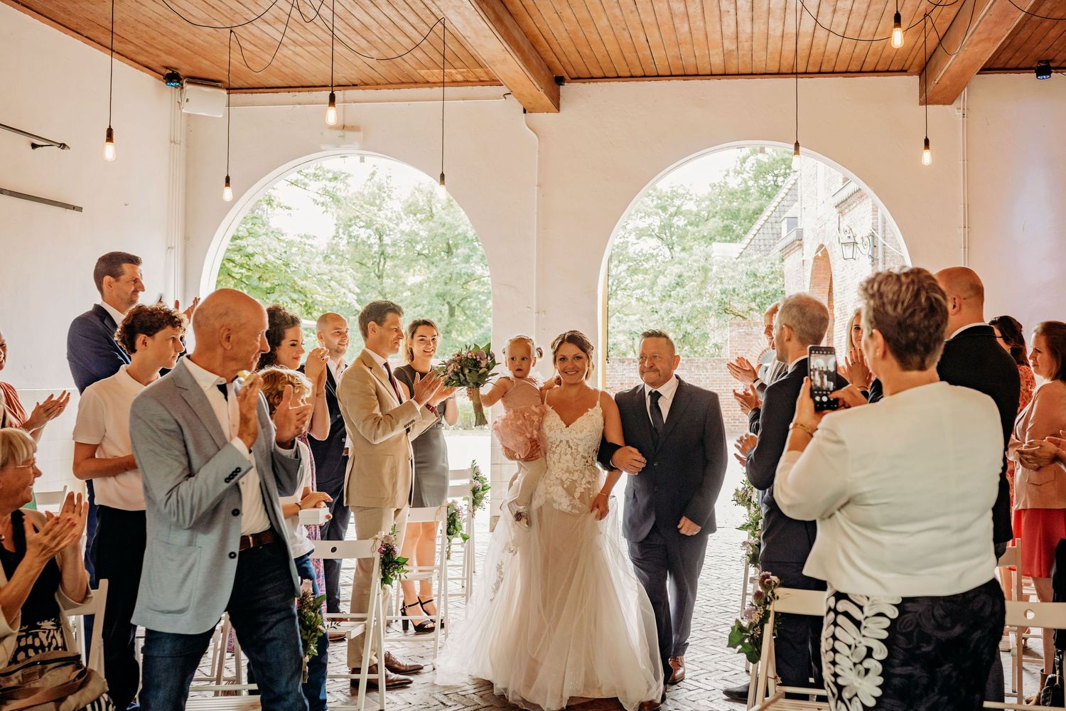 Bride walks down the aisle with guests celebrating at a bright white wedding venue.