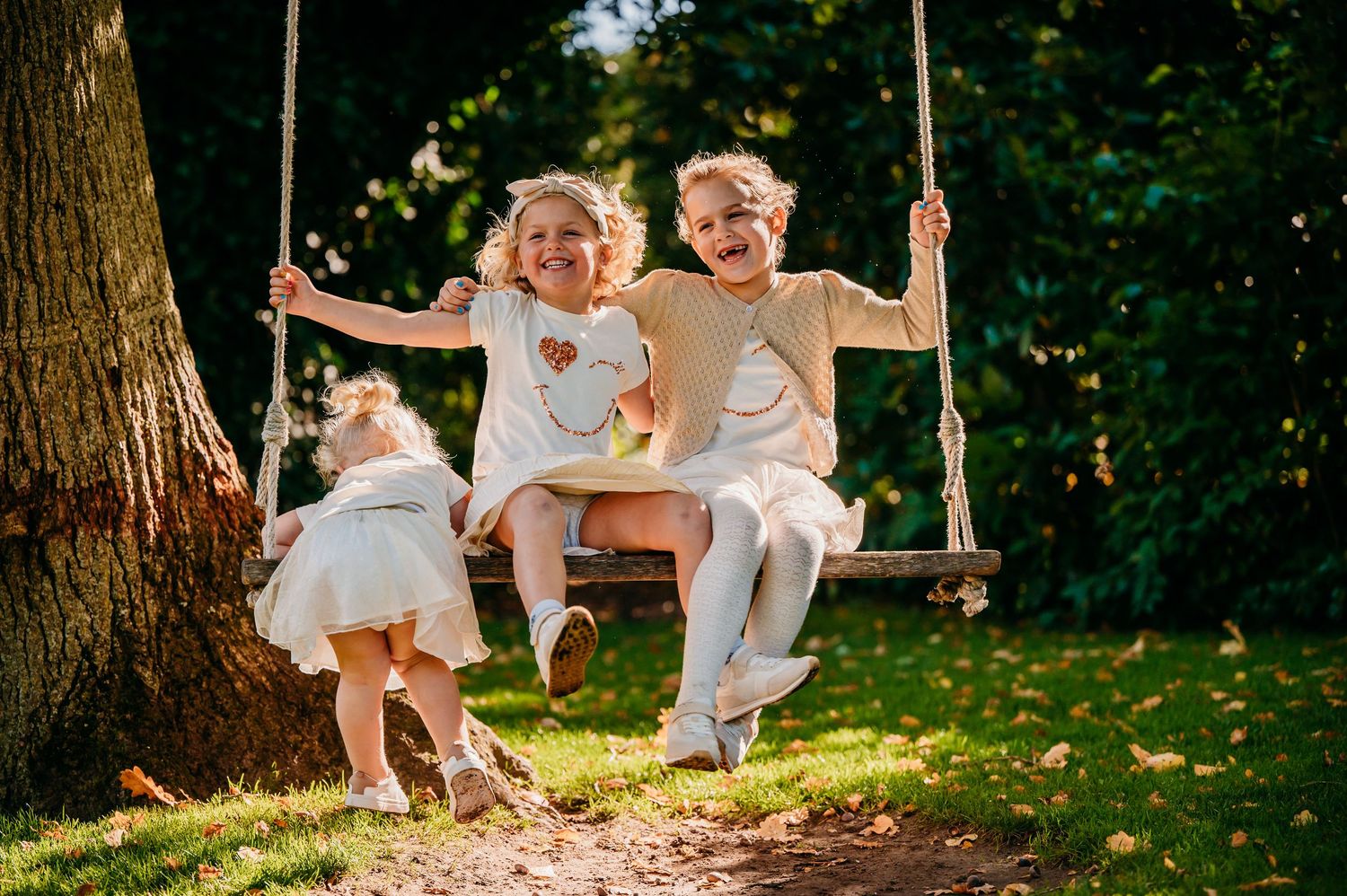 Drie kinderen in witte kleding spelen op een houten schommels tijdens zomerbruiloft, trouwfotografie Brabant.