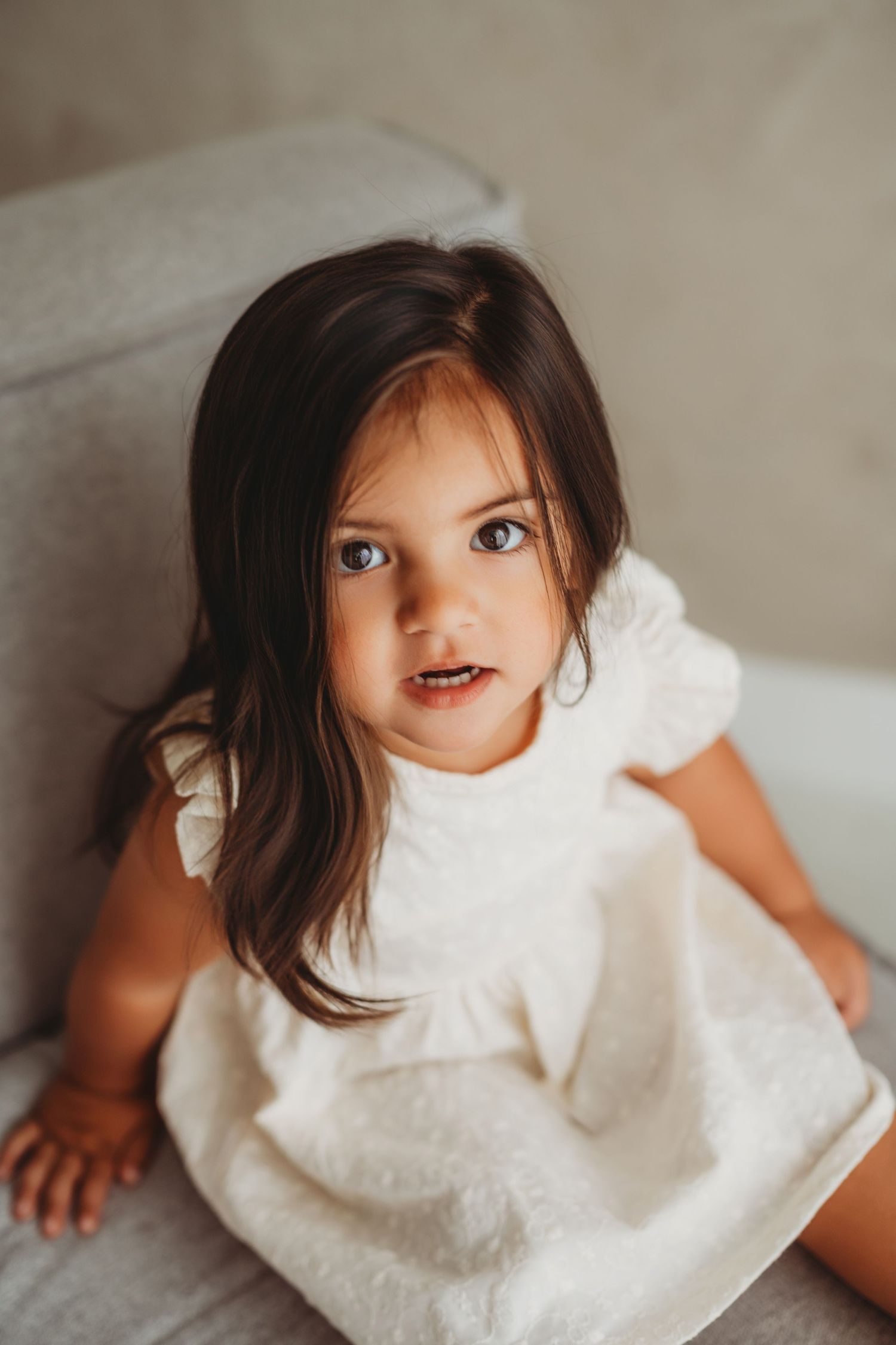 A young girl with dark hair wearing a white ruffled dress sits on a gray sofa, looking up with big blue eyes.