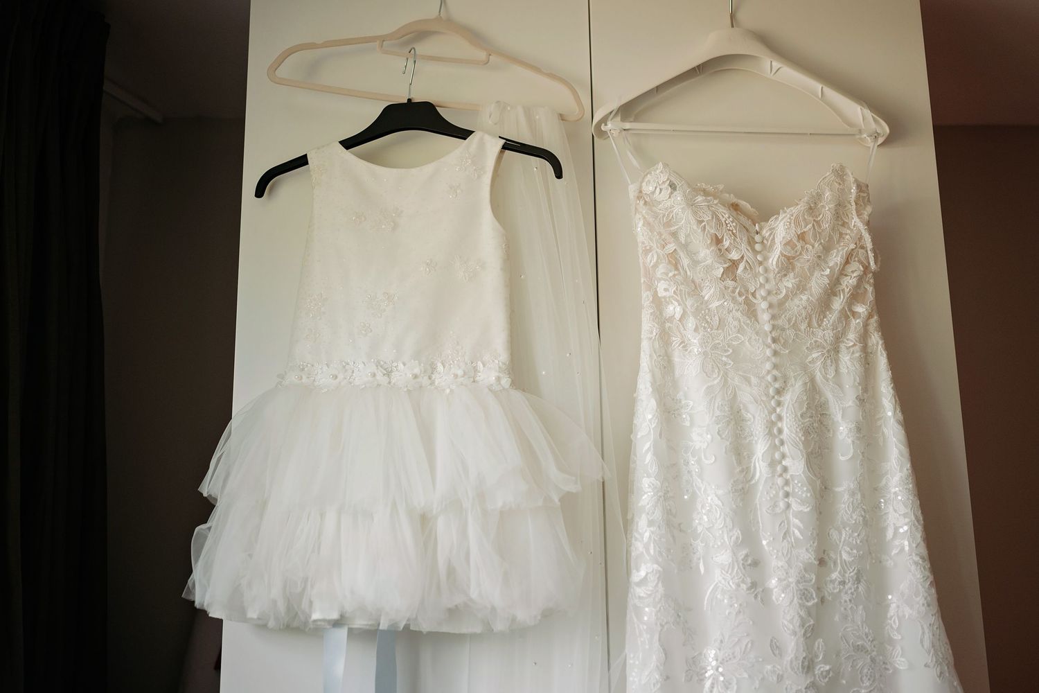 Two white wedding dresses hanging on a closet door, including a flower girl dress and a lace bridal gown.