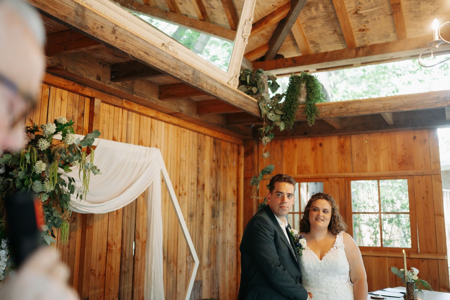 Bride and groom stand at rustic wooden chapel altar decorated with greenery and floral arrangements during wedding ceremony.