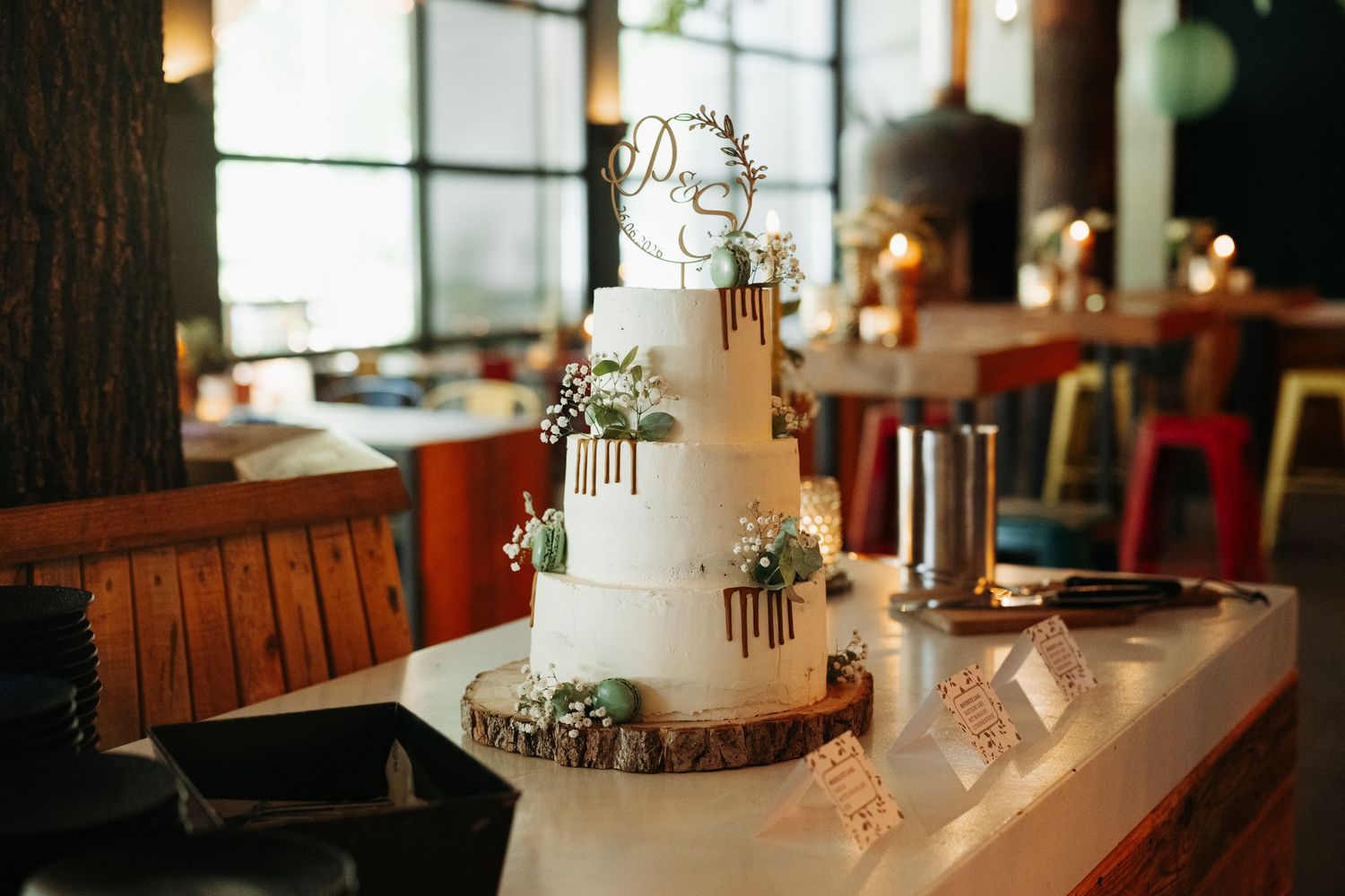 Three-tier white wedding cake with gold drip and floral decor on rustic wood slice stand.