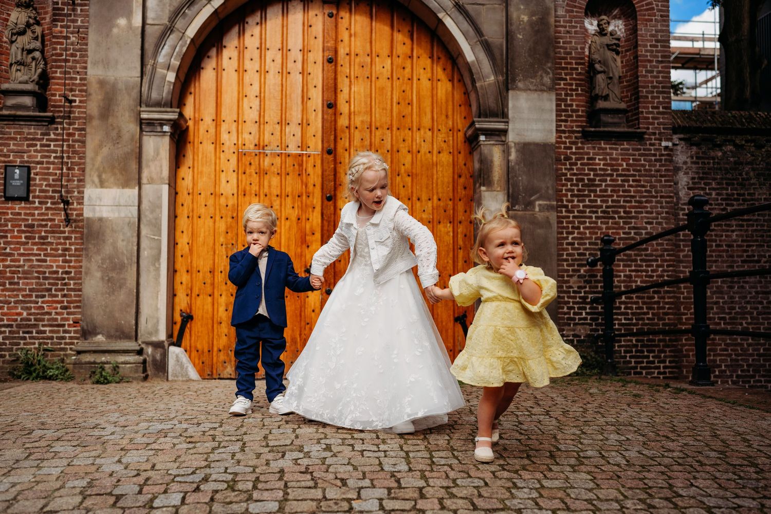 A flower girl in white dress holds hands with two children in blue suit and yellow dress outside a brick church.