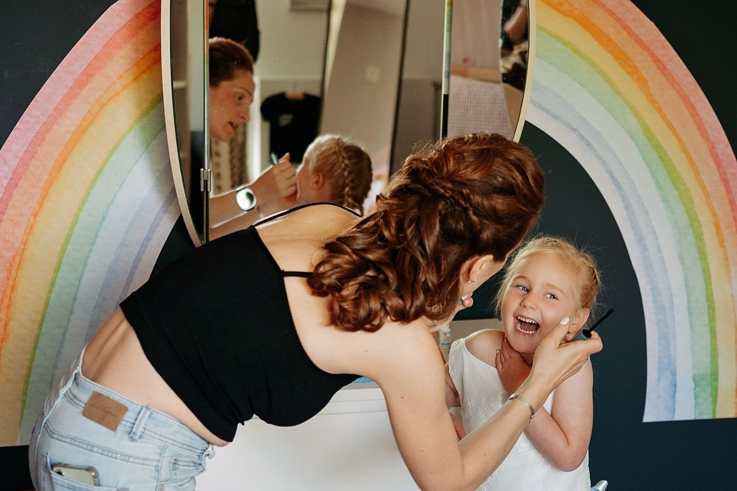 Woman leans toward laughing girl in front of rainbow mirror, sharing a joyful moment together.