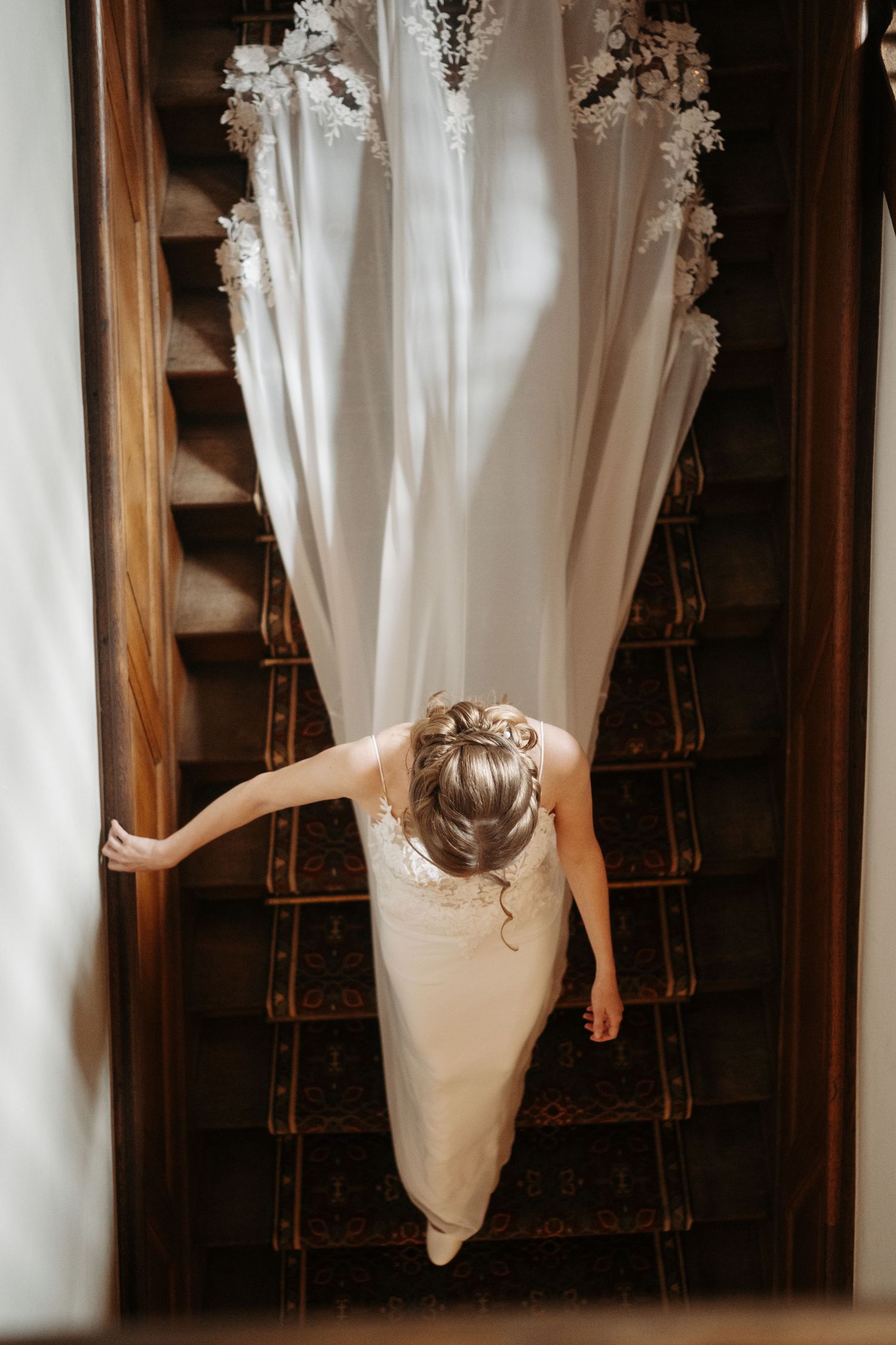 Aerial view of a bride in a white gown descending a wooden staircase with a flowing veil.