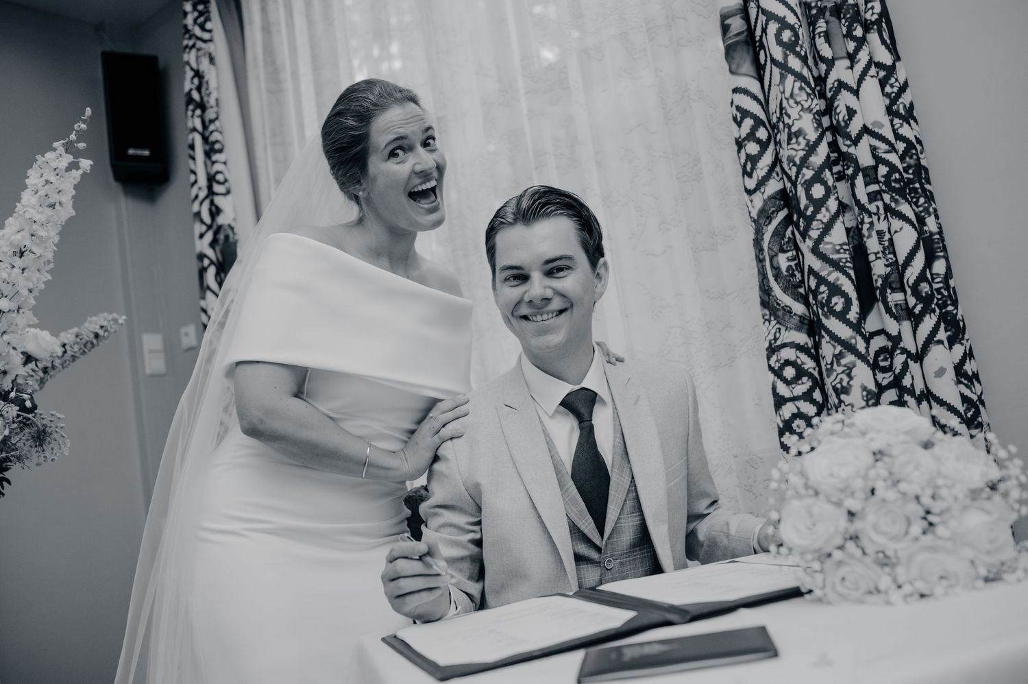 Black and white photo of a bride and groom laughing joyfully while signing the wedding register.