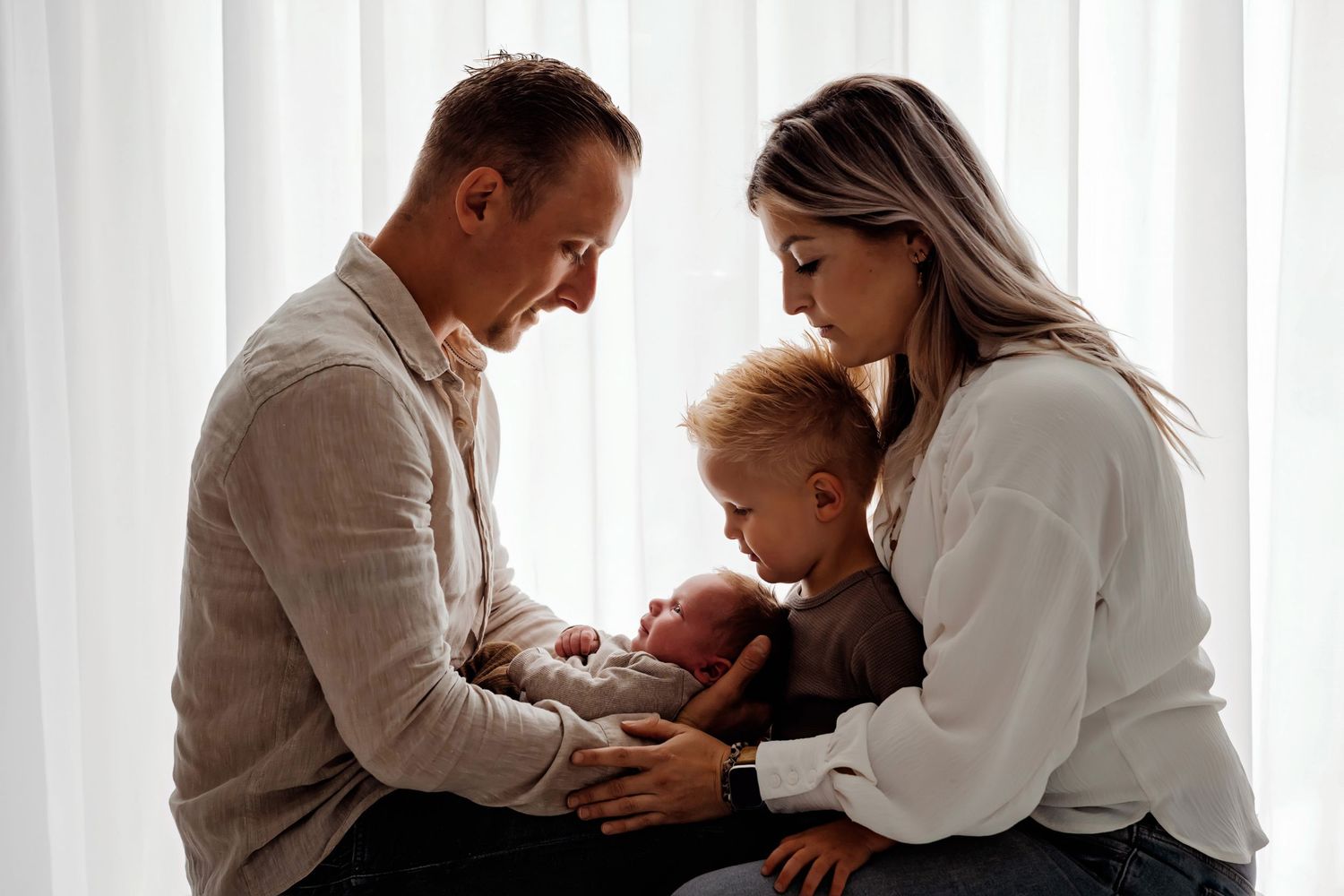 Familie deelt intiem moment bij helder raam met witte gordijnen, familieshoot Brabant