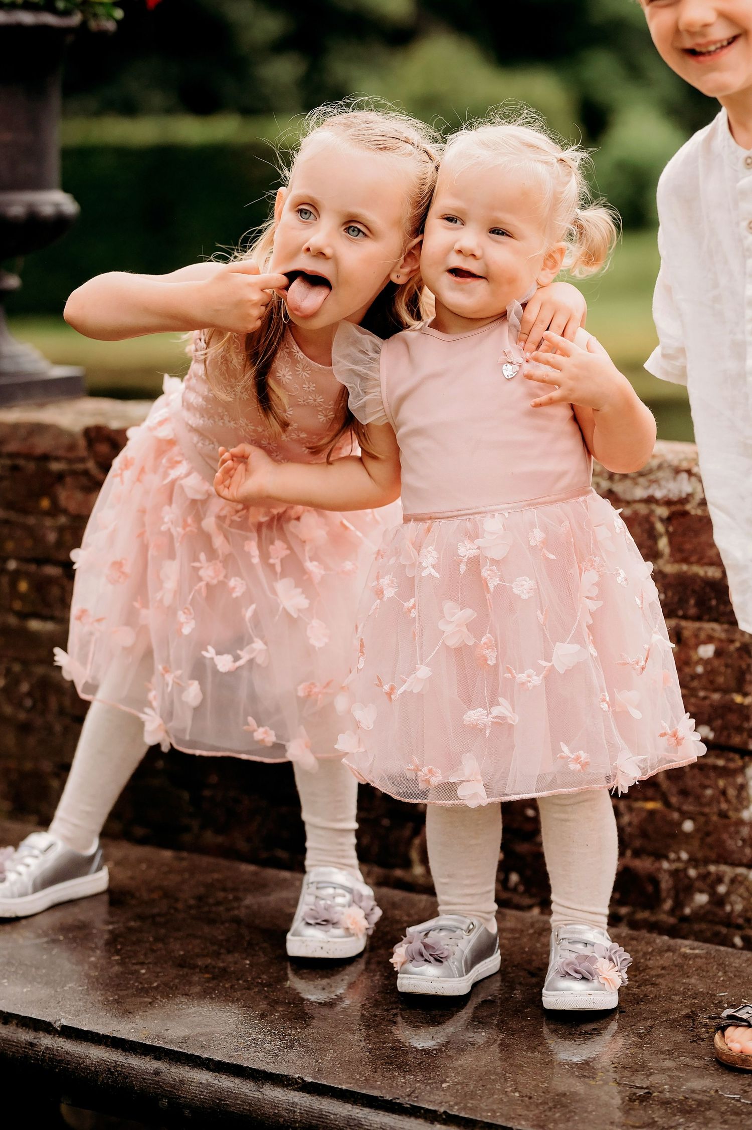 Two adorable little girls in matching pink tulle dresses hugging and making funny faces at a wedding.