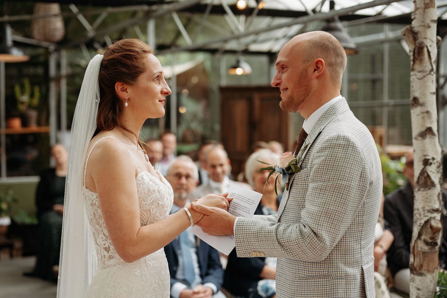 Bride and groom exchanging vows at indoor wedding ceremony surrounded by guests in greenhouse venue.