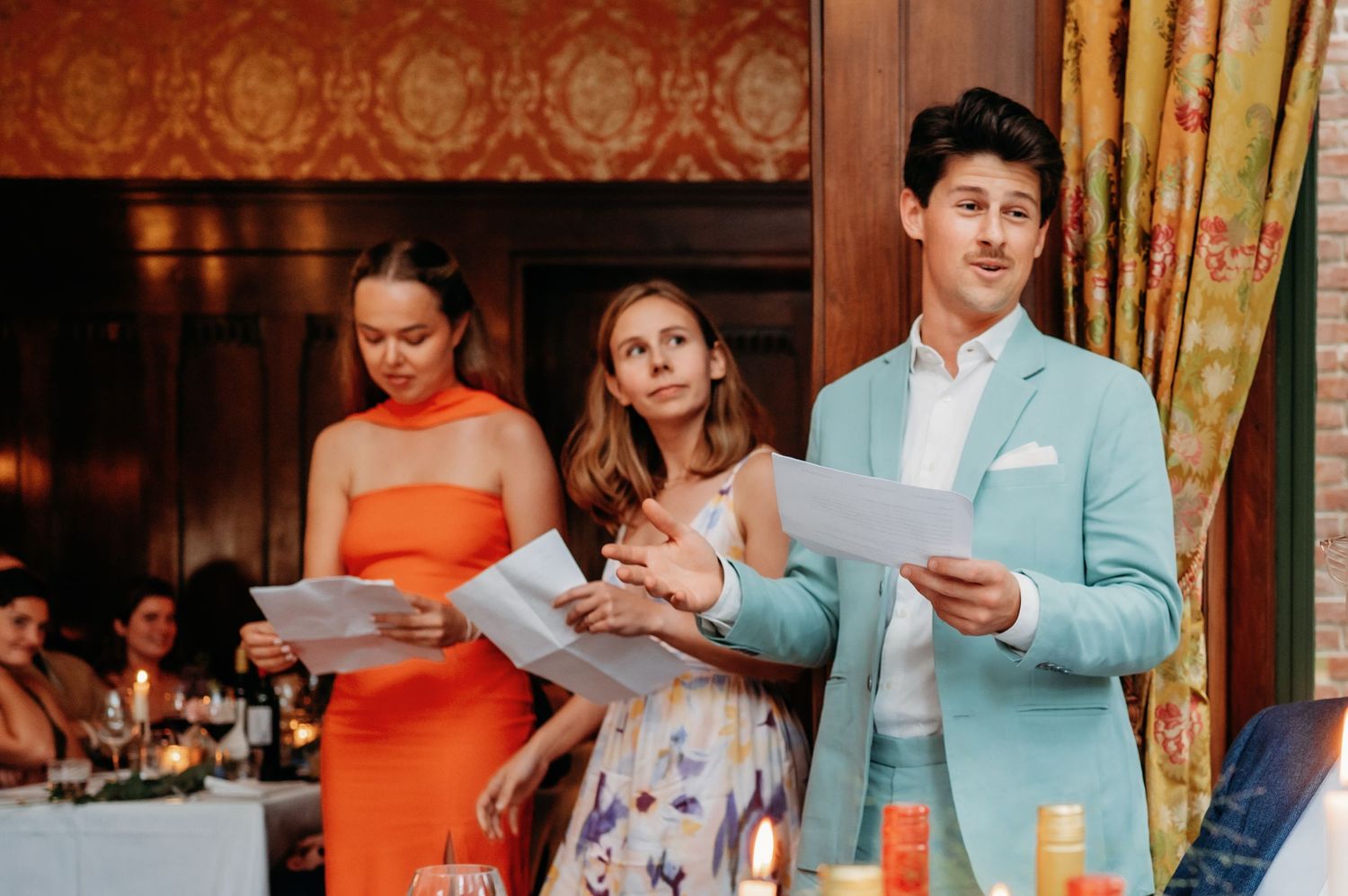 Three guests giving a speech at a wedding reception, with two women and a man in a light blue suit holding papers.