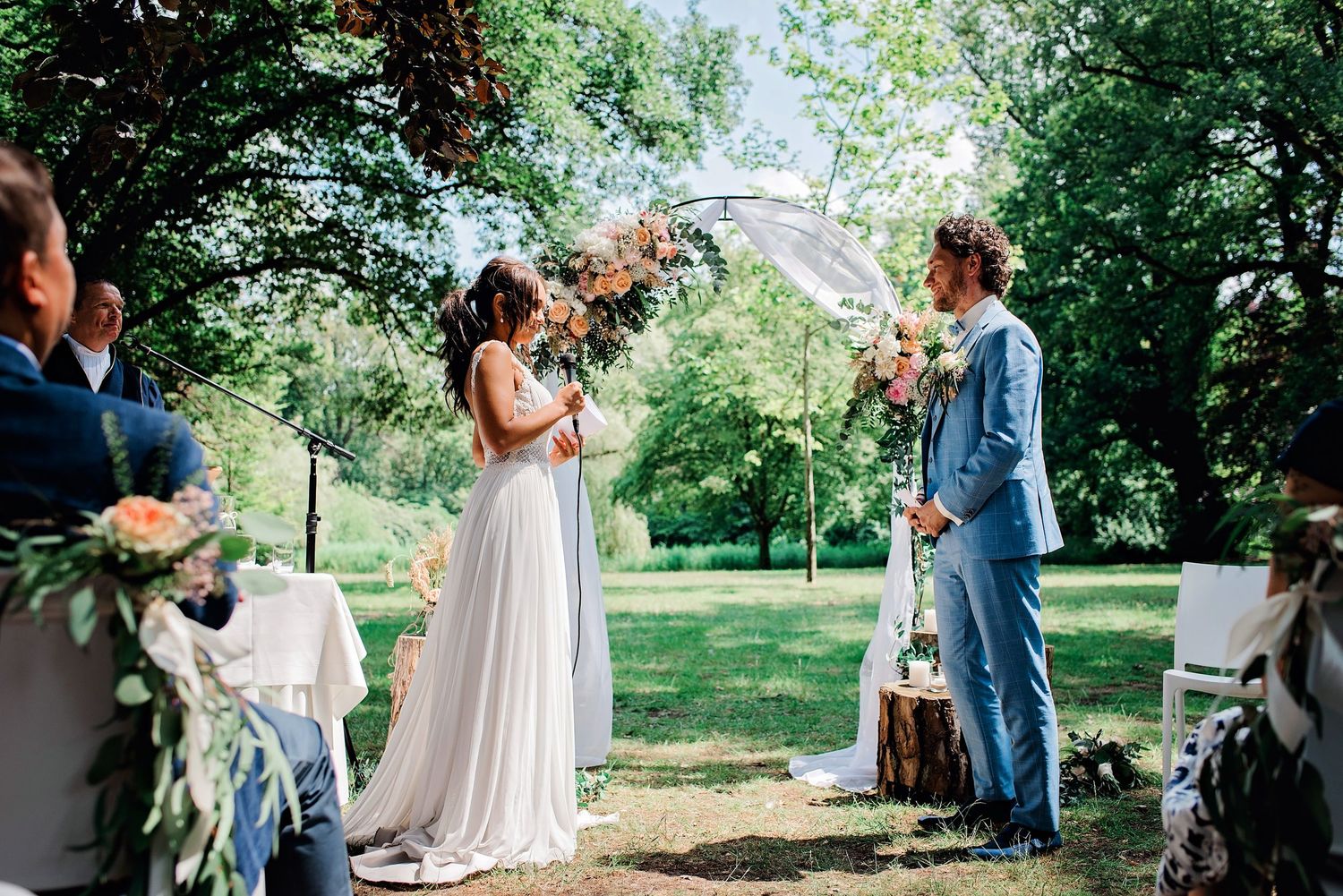 Wedding ceremony setup outdoors with chairs arranged under tall trees in a garden.