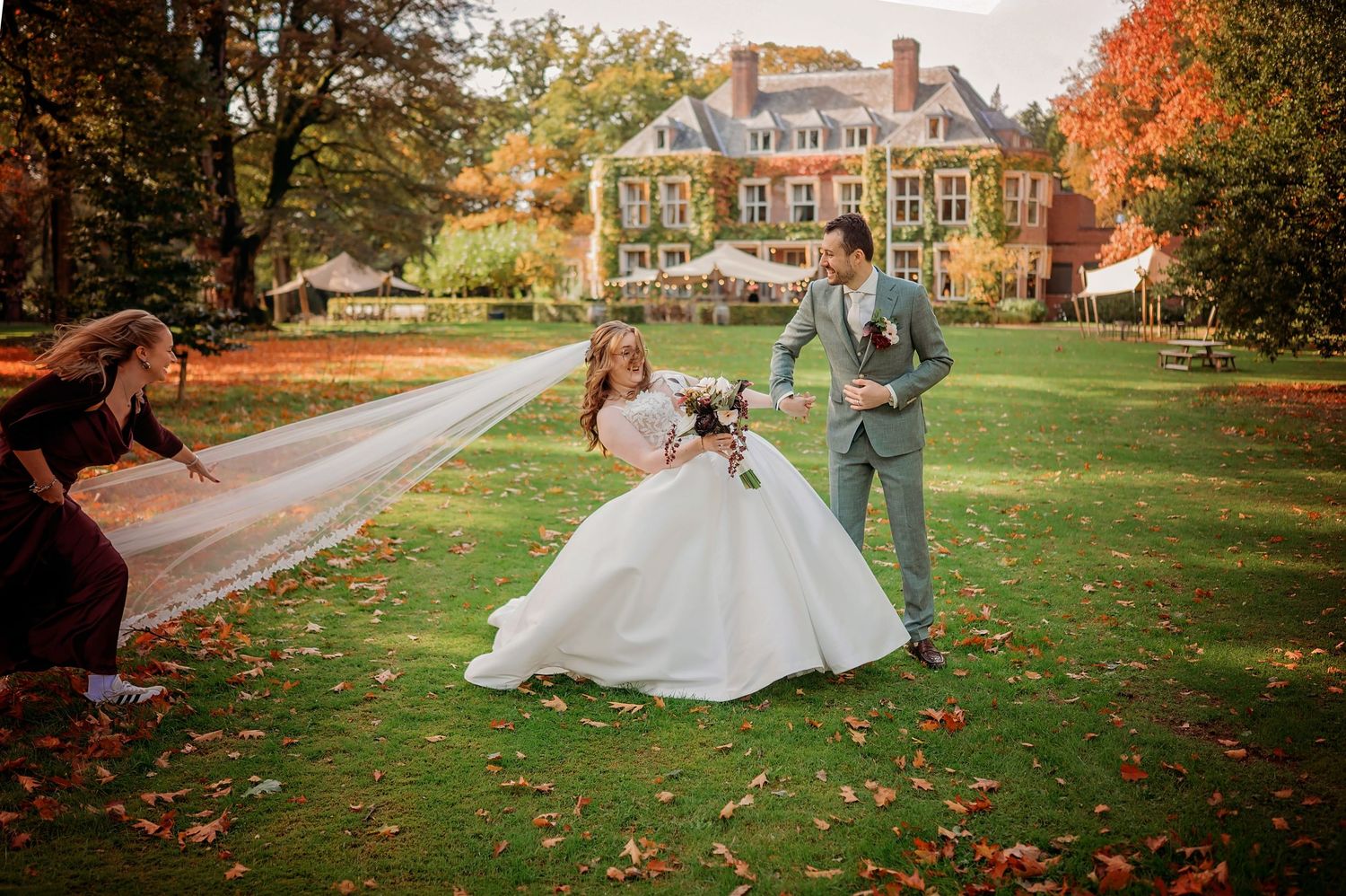 Bride in a white gown and groom in a gray suit pose on an autumn lawn as a woman holds her long veil outstretched before a manor house.