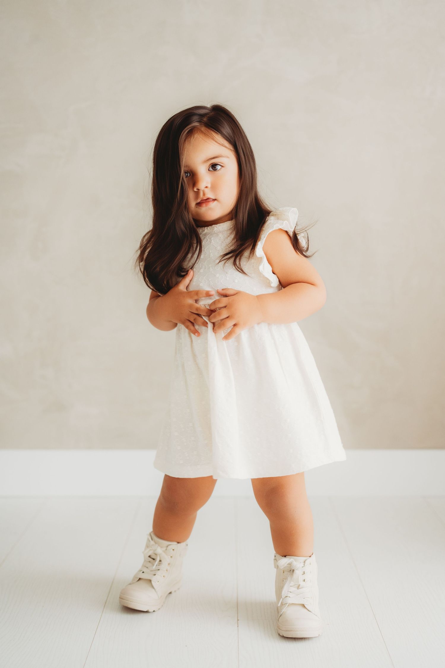 A toddler girl in a white dress and sneakers with long dark hair poses against a light gray wall.
