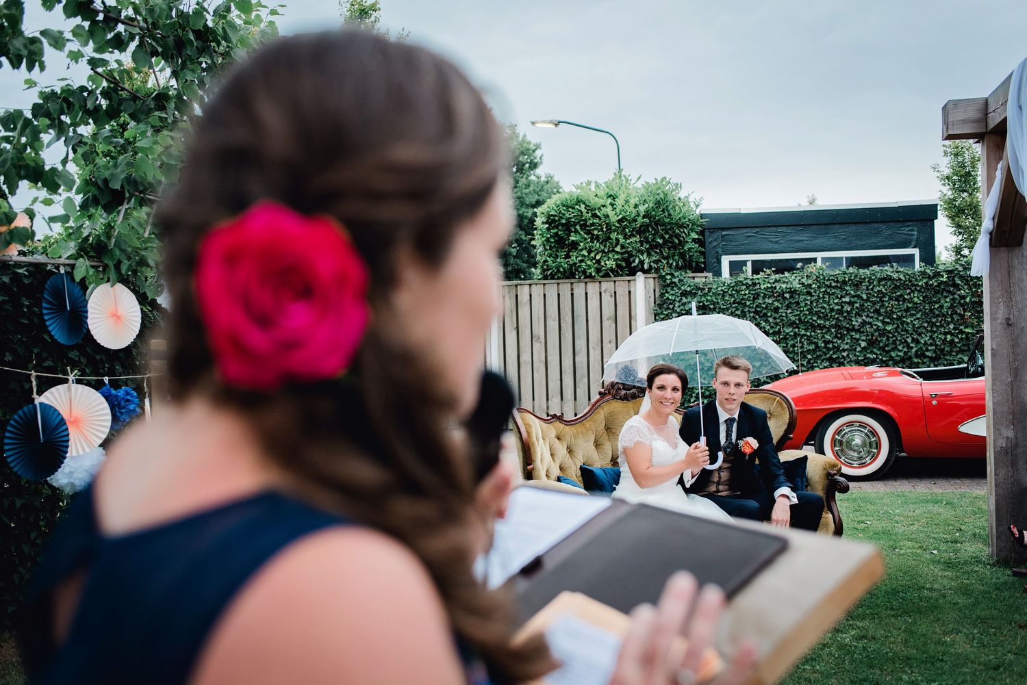 Buitenceremonie met klassieke rode auto en paraplu op achtergrond, trouwfotografie Brabant.