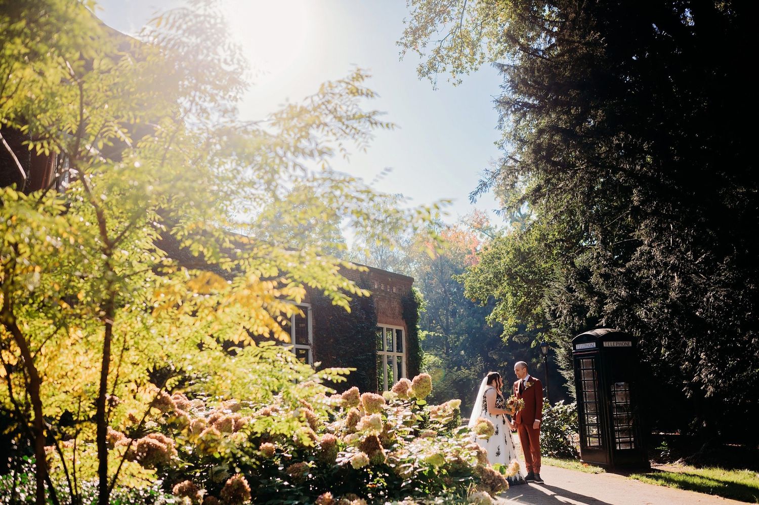 Bruidspaar loopt over zonovergoten tuinpad met herfstlicht door bomen, bruidsfotografie Huize Bergen Vught, Brabant.