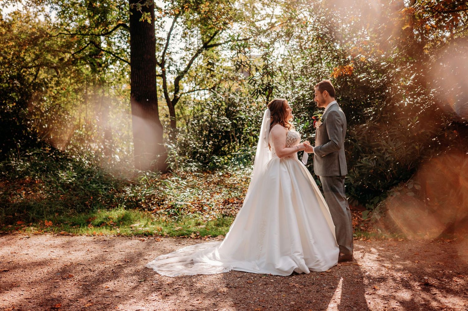 A bride in a white ball gown and veil stands with her groom in a gray suit on a sunlit forest path in autumn.