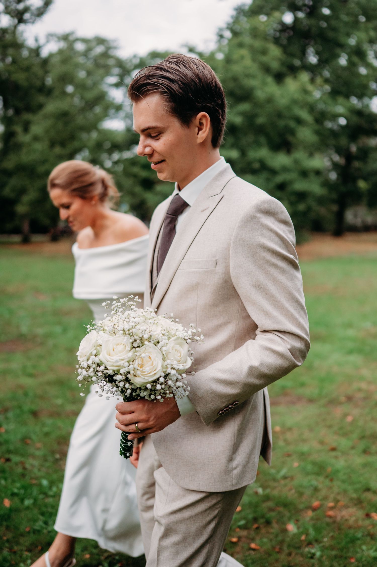Groom in beige suit holds white rose bouquet with baby's breath, bride in off-shoulder dress stands behind in park.