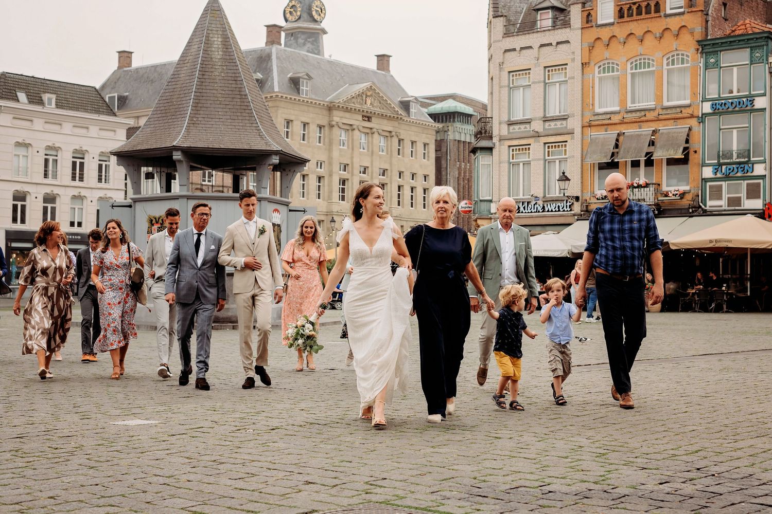 Wedding party walking across a historic European town square, bride in white gown holding bouquet with guests.