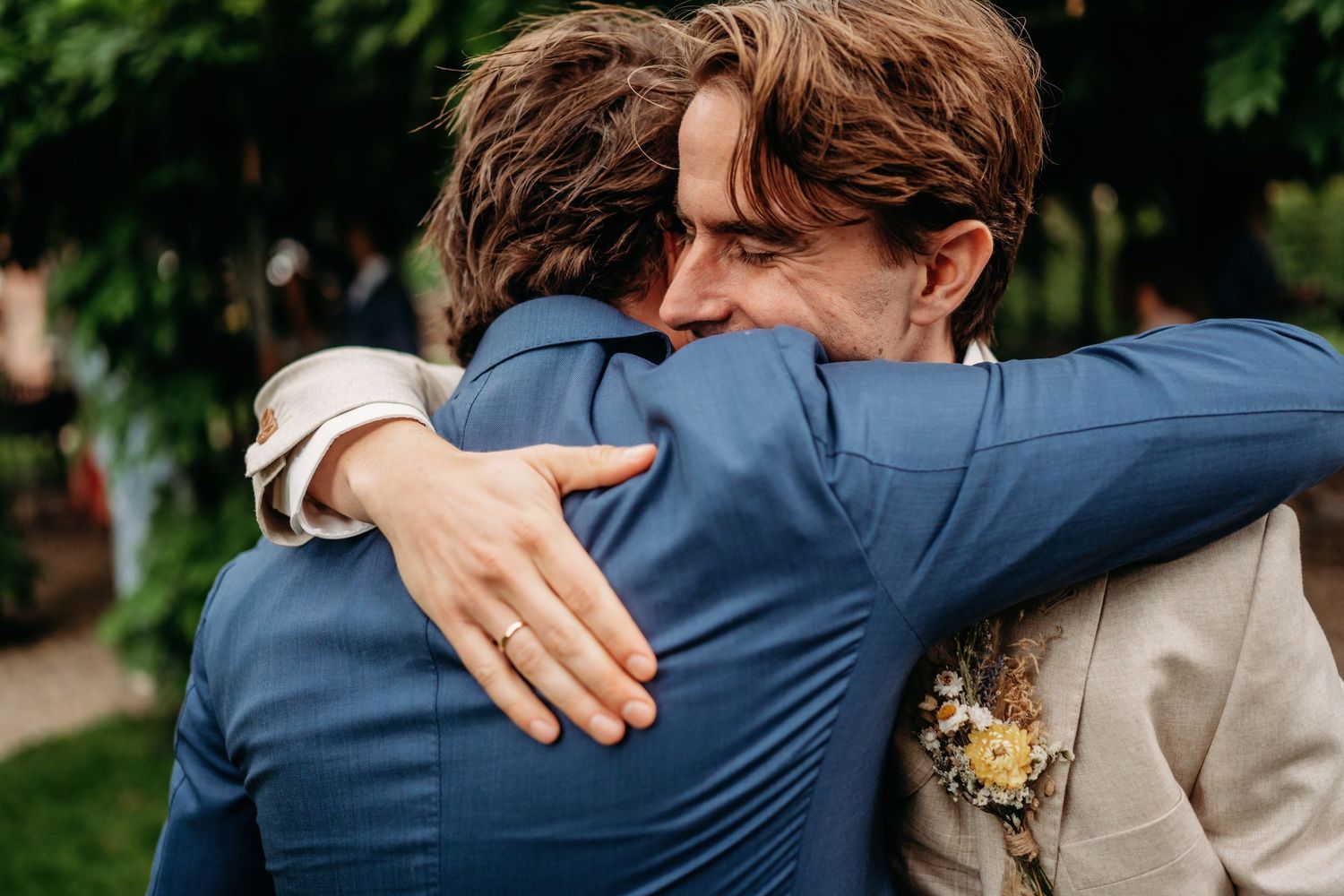 Two men in suits share a warm embrace at a wedding, one wearing a ring and boutonniere.