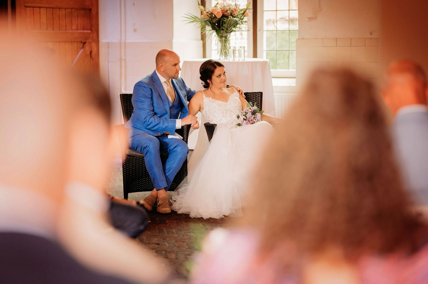 Bride and groom seated together during wedding ceremony, groom in blue suit, bride in white gown, guests in foreground.