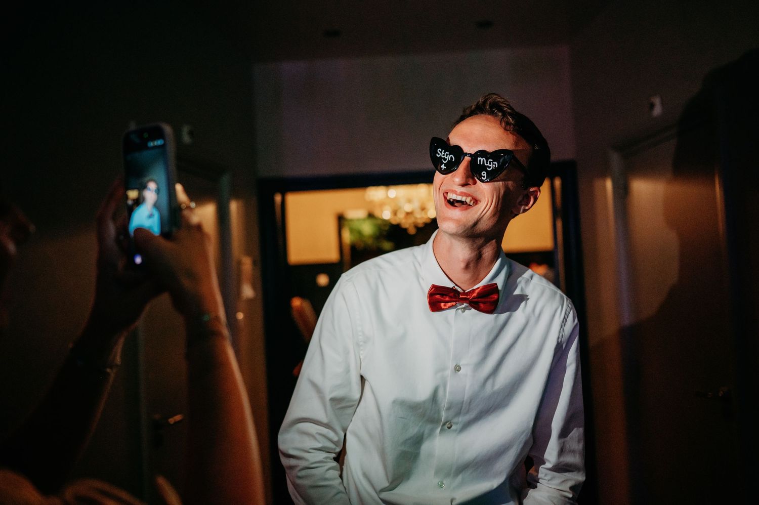 Groom in white shirt and red bow tie wearing novelty sunglasses celebrates at wedding reception.
