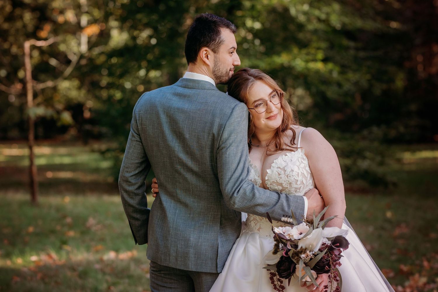 Groom in a gray suit embraces a smiling bride in a white lace gown holding a floral bouquet in an outdoor wedding portrait.