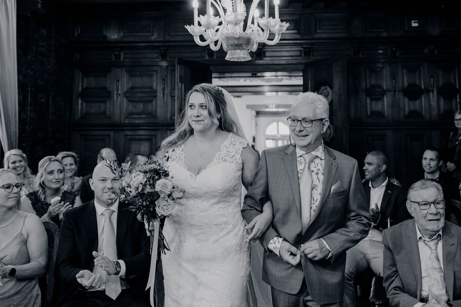 Bride walks down aisle with older gentleman in black and white wedding ceremony photo inside elegant wood-paneled venue.