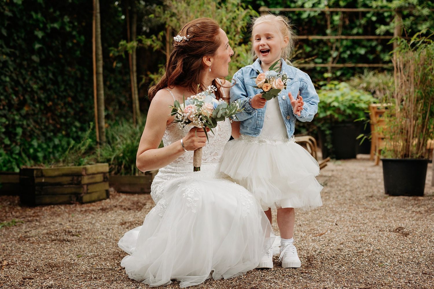 Bride kneeling in white gown holding bouquet, laughing with flower girl in tutu and denim jacket outdoors.