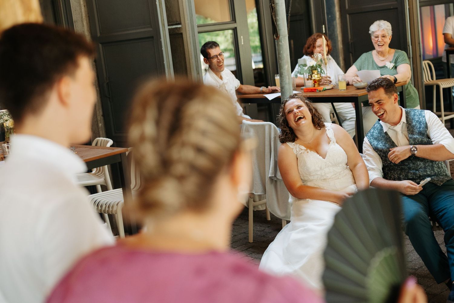 Bride and groom laughing joyfully during wedding reception with guests seated at tables in elegant venue.