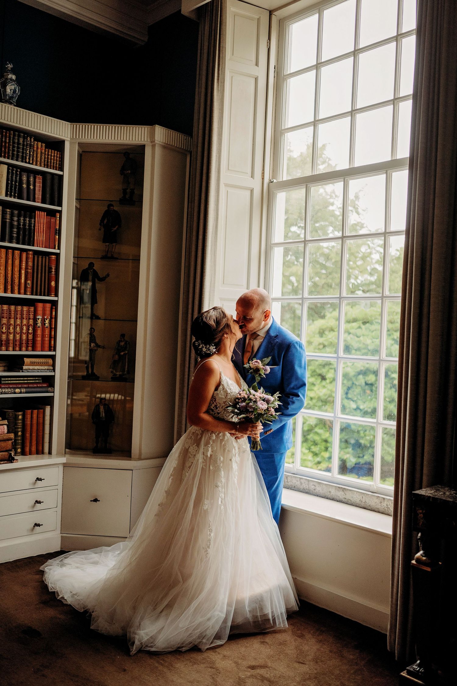 Bride and groom share a romantic kiss by a large window in an elegant library room, bride holding a bouquet.