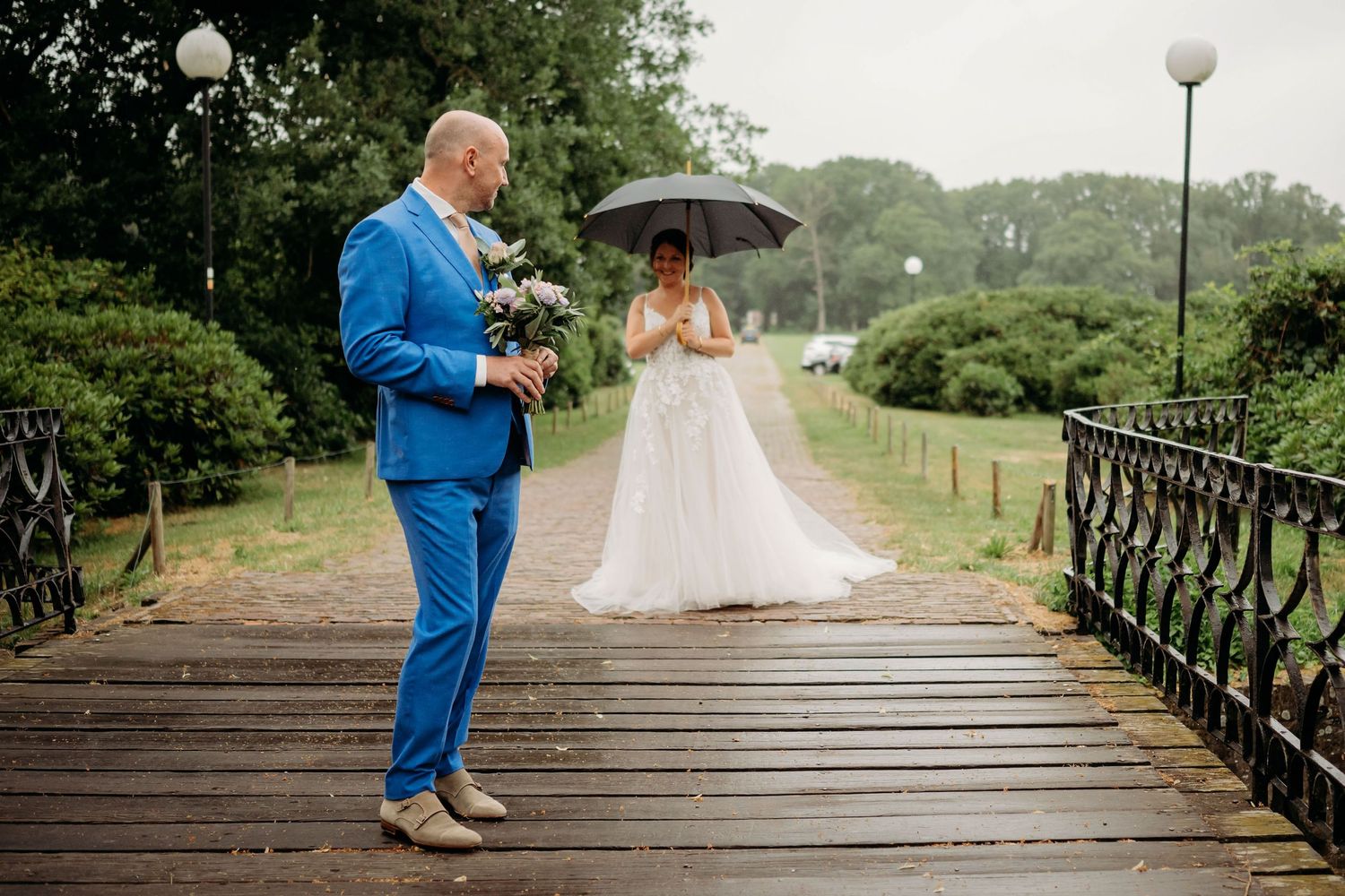 Bride and groom on a rainy wedding day on a wooden bridge, bride holding umbrella in white gown, groom in blue suit.