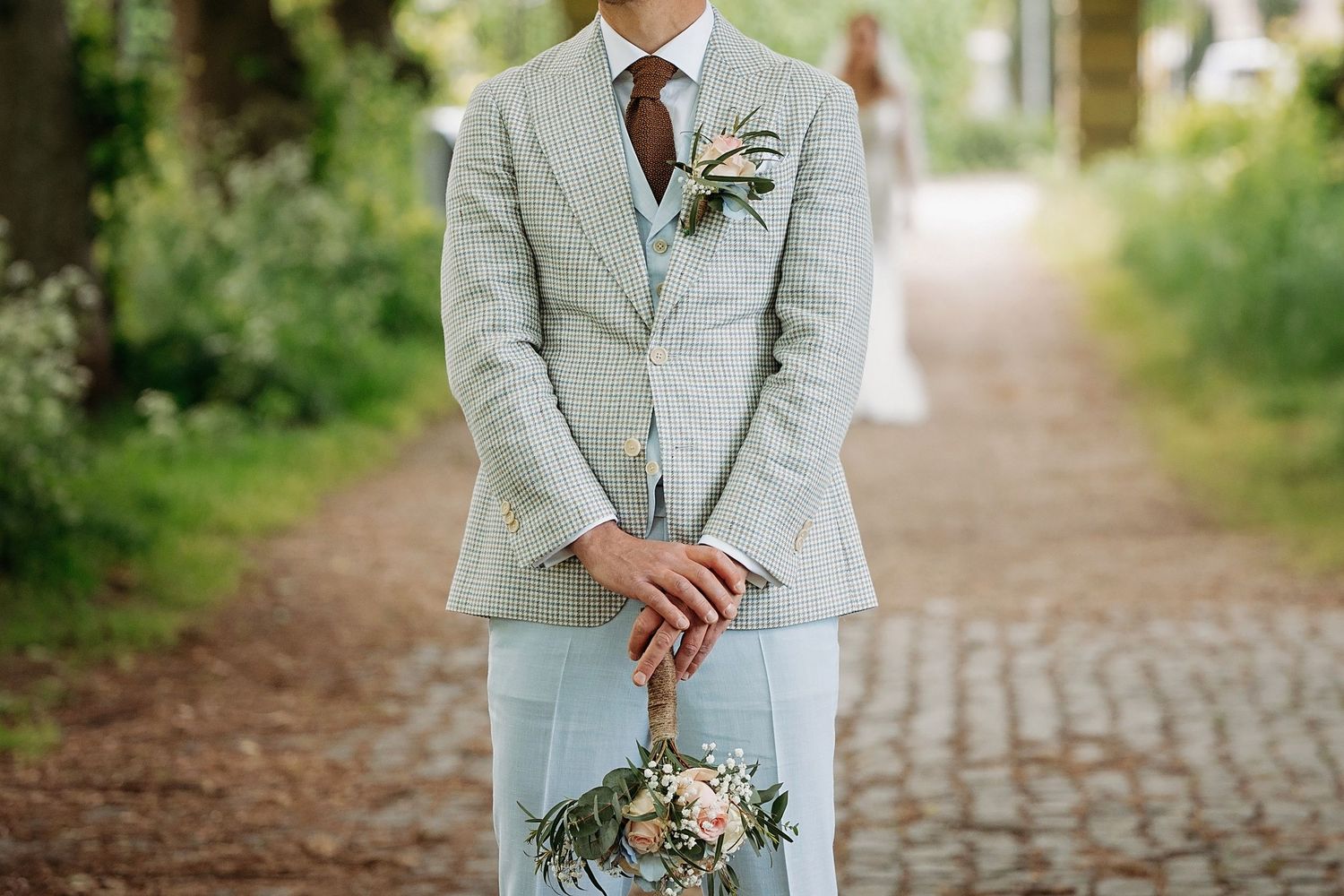 Groom in light gray pinstripe suit holding a bridal bouquet with pink roses and greenery, standing on a brick path.