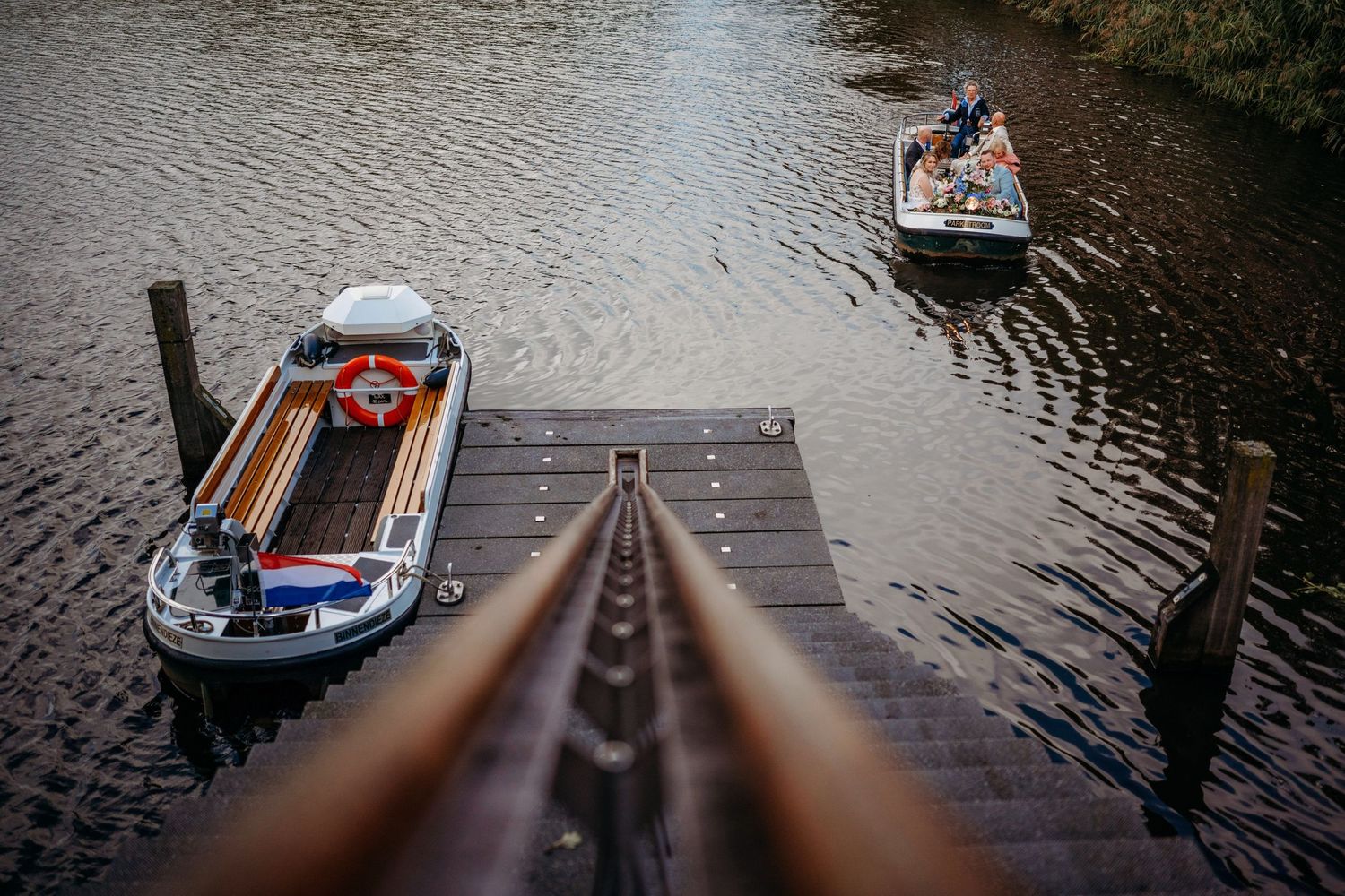 Two boats docked at a wooden pier on a calm river, with a second boat carrying passengers in the background.