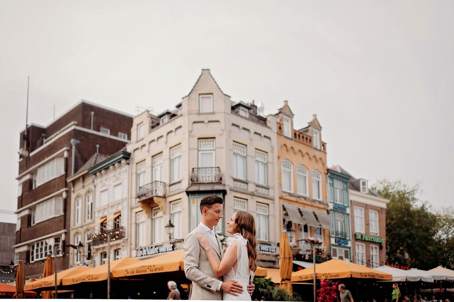 Couple embracing in front of colorful European buildings at an outdoor market square on a cloudy day.
